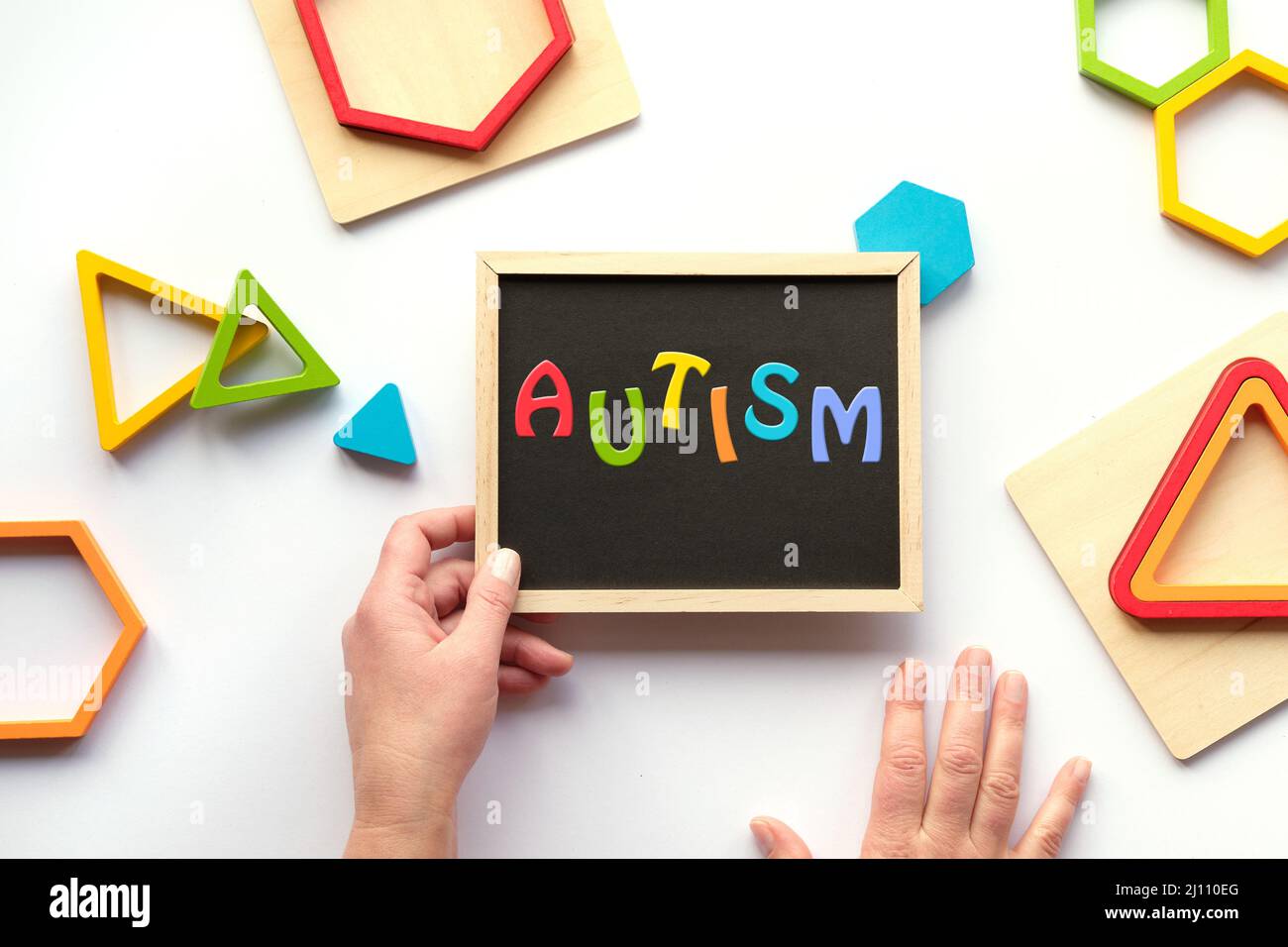 Text Autismus, Wort auf Tafel in Regenbogenfarben. Genestete Holz-Dreiecke und Sechsecke in verschiedenen kräftigen Farben. Kreidetafel in menschlicher Hand. Stockfoto