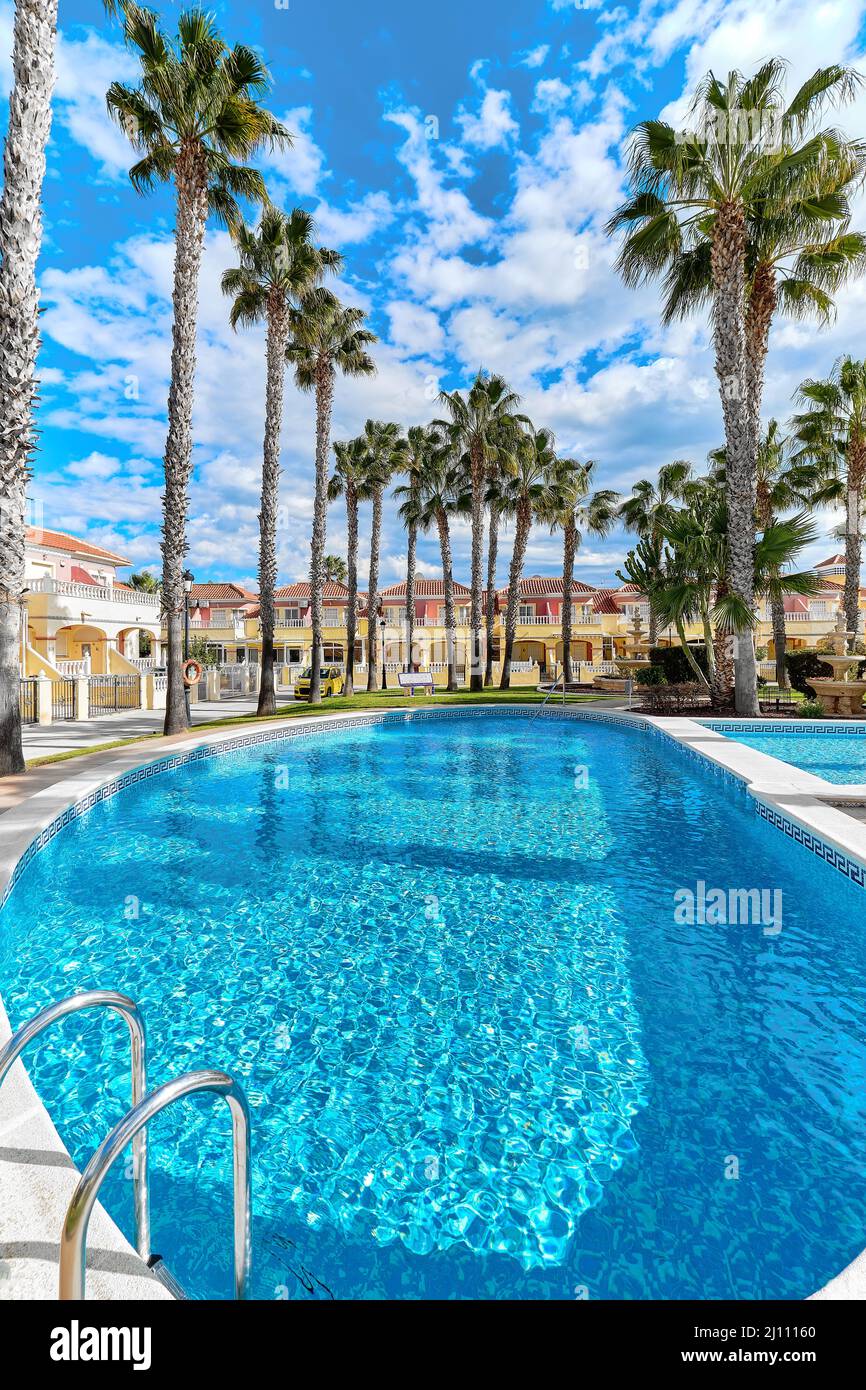 Vertikales Bild Palme um leeres Schwimmbad mit blauem Wasser in Wohnsiedlung mit modernen Luxushäusern gegen bewölkten Himmel bei su Stockfoto