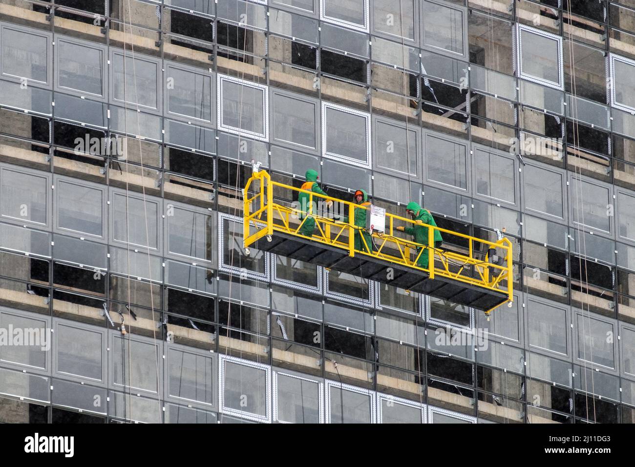 Die Arbeiter in der Bauwiege in der Höhe sind im Winter mit der Verglasung der Fassade eines Wolkenkratzers beschäftigt. Side Vie Stockfoto