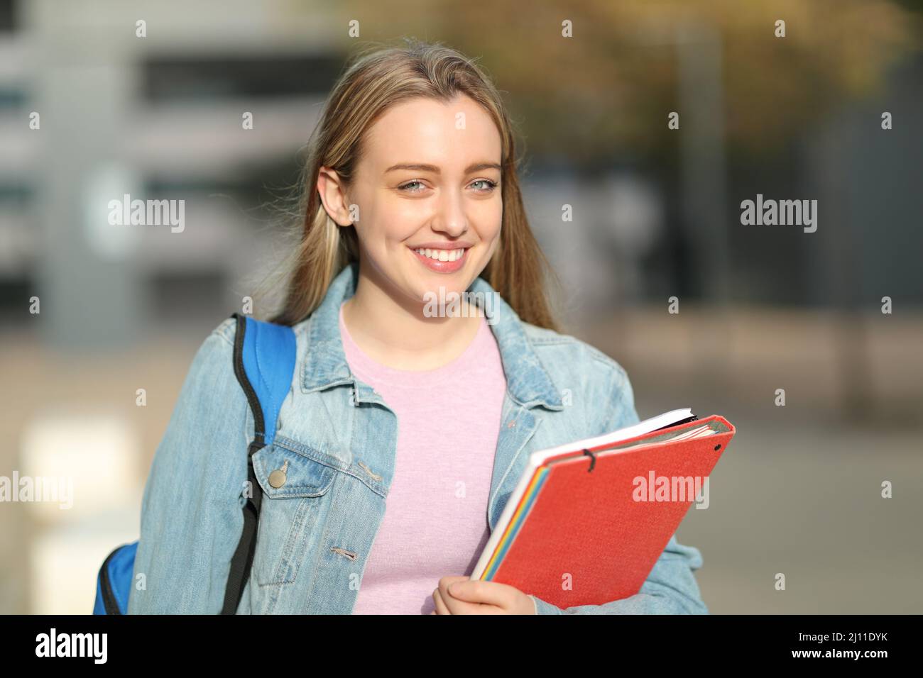Fröhlicher Student, der Ordner und einen Rucksack in der Hand hält und die Kamera auf einem Campus anschaut Stockfoto
