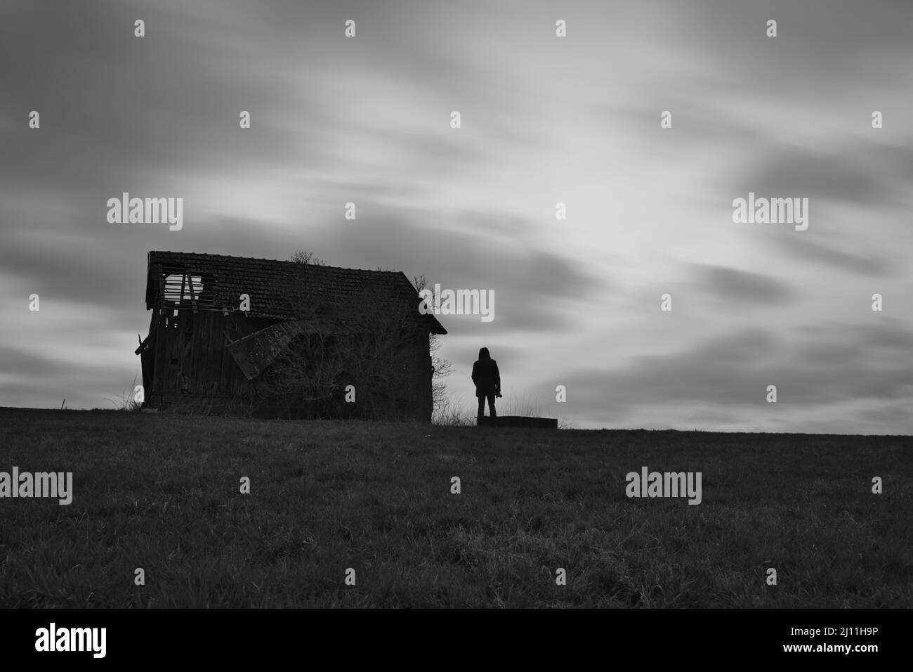 Gruselig verlassene Scheune auf einer Wiese mit Wolken im Hintergrund und einer Silhouette eines Mannes mit Kamera Stockfoto