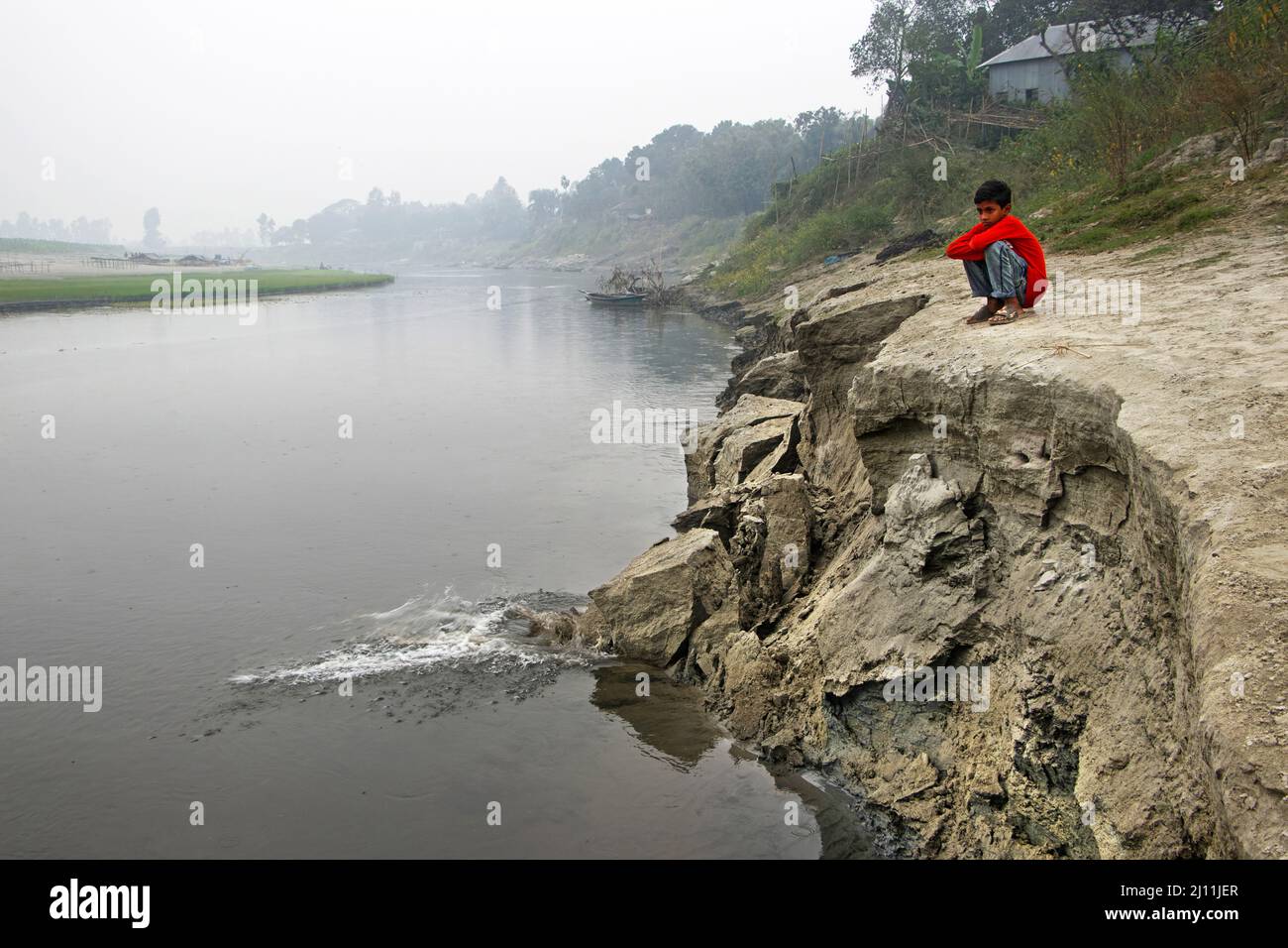 Kamrul, 11, saß am Brahmaputra River in Manikganj, Bangladesh, neben einem erodierenden Flussufer, mit Tränen beäugtem Blick, als die Erosion weiter stattfindet und seine Familie dem Risiko einer Vertreibung aussetzt. Kamruls Familie lebt neben dem Brahmaputra Fluss, muss sich aber nach unbekannten Zielen aufmachen, nachdem die Erosion durch den Fluss den größten Teil ihres Landes und ihrer Heimat weggenommen hat. Bangladesch mit seiner dichten Bevölkerung und seiner Fülle an Flüssen hat mehr als seinen Anteil an tropischen Stürmen, Überschwemmungen und anderen Naturkatastrophen überstanden. Der Klimawandel hat diese zerstörerischen Elemente nur beschleunigt. Aber es steht auch einem anderen gegenüber, mehr Stockfoto