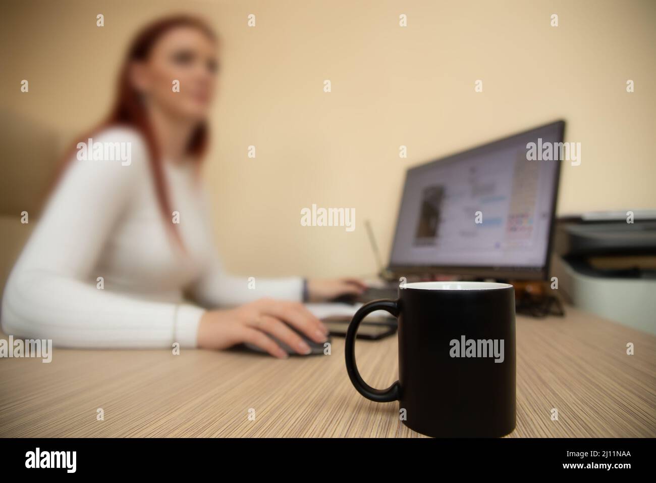 Eine Büroangestellte arbeitet an einem Computer. Im Vordergrund ist ihr schwarzer Kaffeebecher. Stockfoto