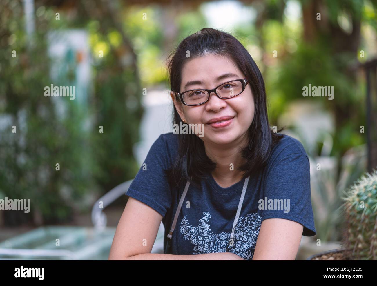 Porträt einer asiatischen Frau mittleren Alters trägt Brillen, kurze schwarze Frisur, sitzt in einem Garten, lächelndes Gesicht, Platz für Text. Stockfoto