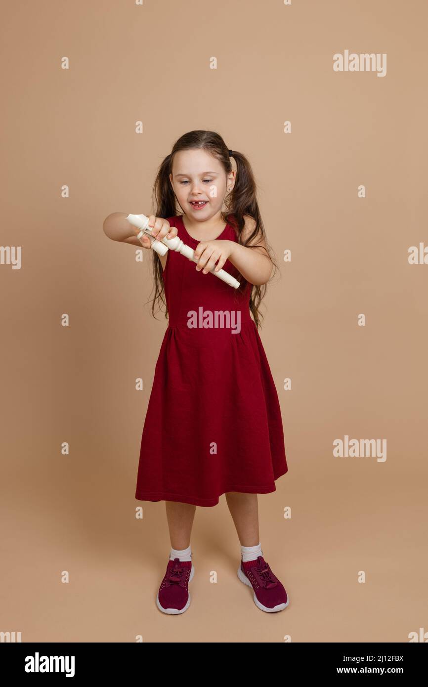 Mädchen in roten Kleid begeistert demontieren gemeinsame Flöte in Teilen, beige Hintergrund. Lernen, Holzbläser-Musikinstrument zu spielen. Stockfoto