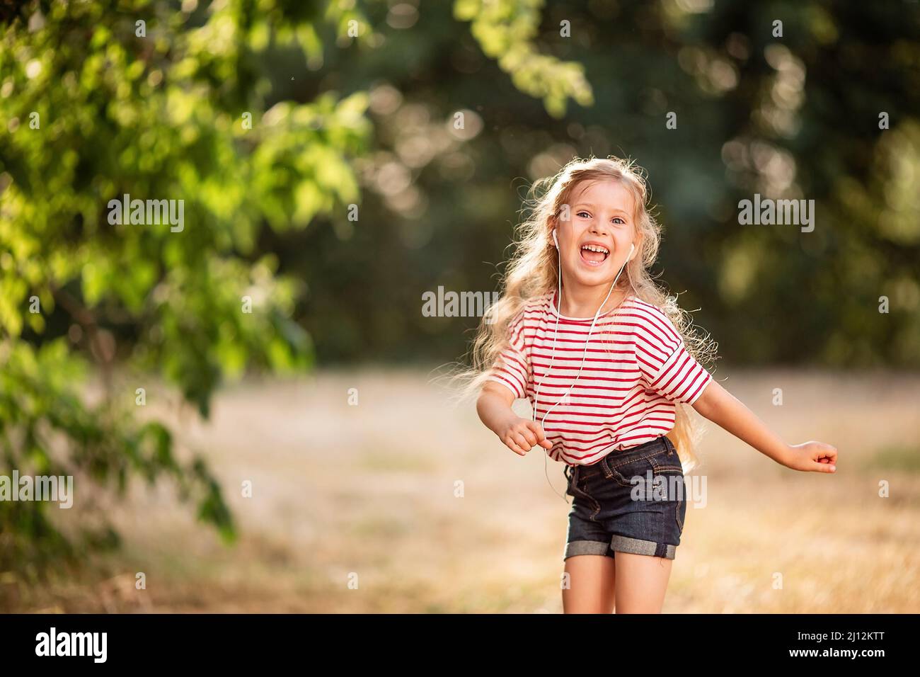 Glückliches kleines blondes Mädchen, das mit Kopfhörern Musik hört, in der Natur im Park tanzt und singt. Stockfoto