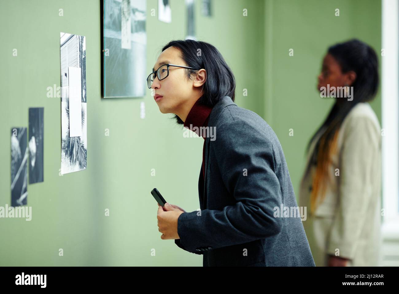 Porträt eines hübschen jungen asiatischen Mannes mit Brille, der eine Ausstellung in einer Kunstgalerie besucht, die sich auf ein Schwarzweiß-Foto an der Wand konzentriert Stockfoto