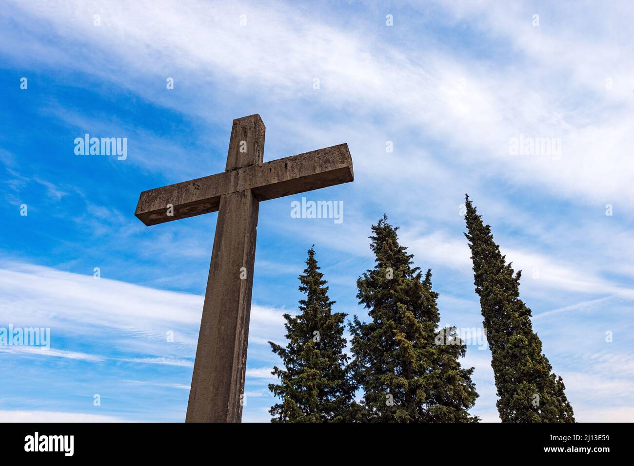 Nahaufnahme eines religiösen Steinkreuzes an einem blauen Himmel mit Wolken, drei Bäumen, zwei Kiefern und einer Zypresse und Kopierraum. Trentino Alto Adige, Italien Stockfoto