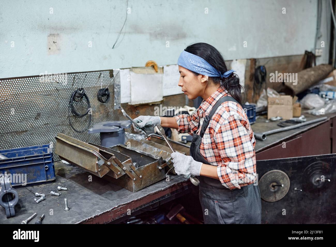 Junge Arbeiterin einer Industrieanlage, die einen riesigen Schraubenschlüssel für Reparaturarbeiten auswählt, während sie an der Werkbank mit einem Metallkoffer steht Stockfoto