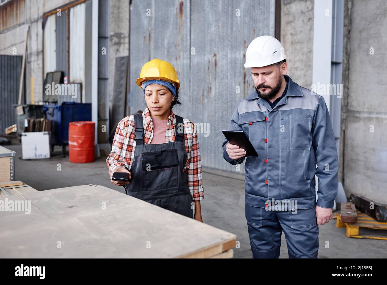 Junge Ingenieurin und Vorarbeiter in Sicherheitshelmen und Arbeitskleidung, die Barcodes auf Kartons mit Ersatzteilen in der Werkstatt prüfen Stockfoto