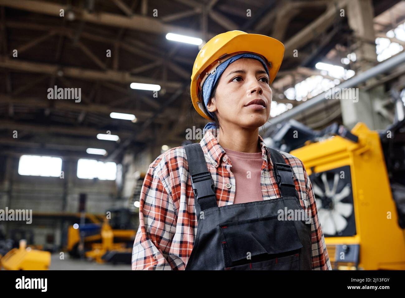 Junge ernsthafte Arbeiterin eines Industriewerks, die Overalls und Hardhut trägt und in einem großen Distributionslager steht Stockfoto