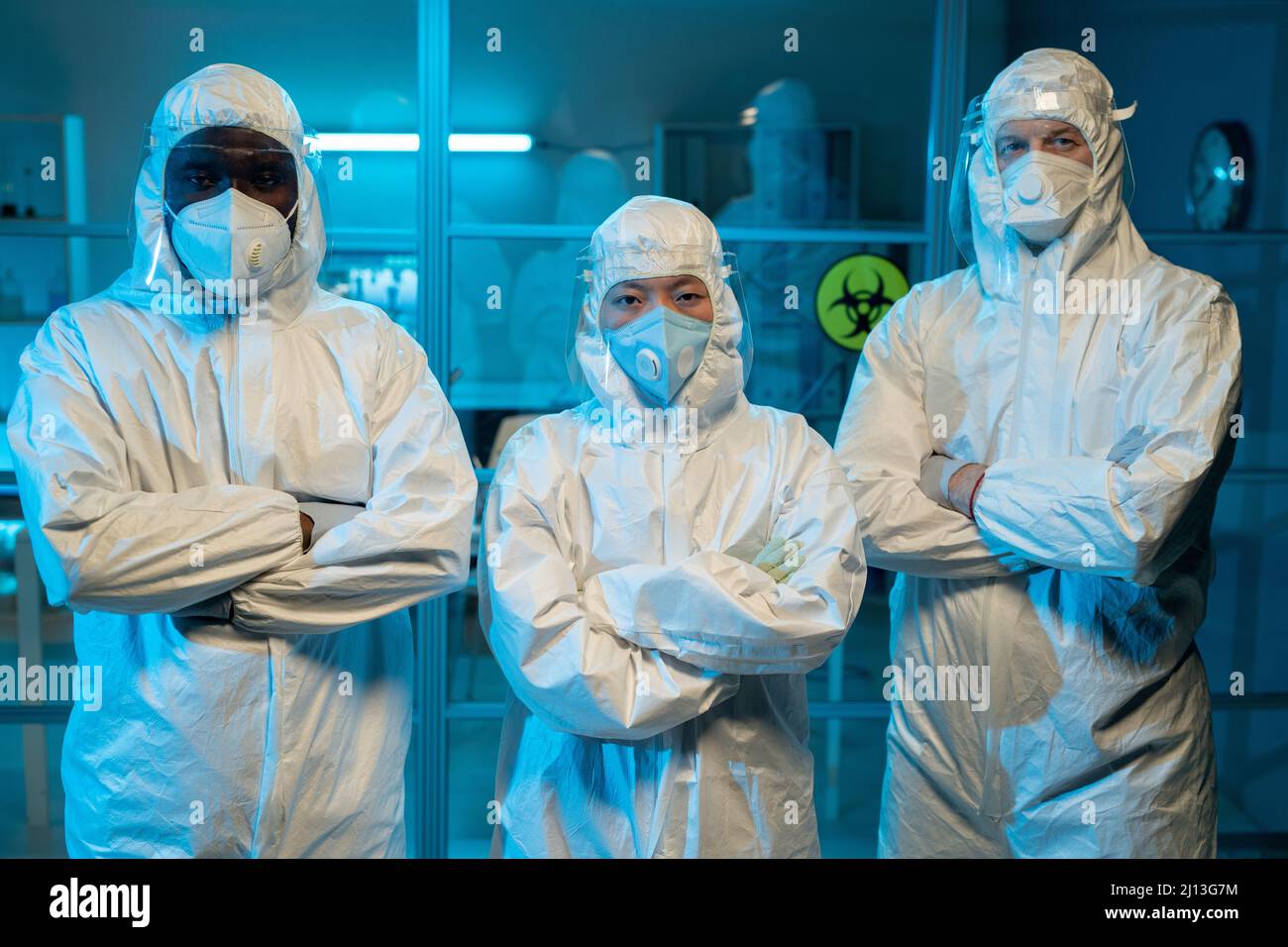 Gruppe zeitgenössischer interrassischer Forscher in schützender Arbeitskleidung, die Arme an Brust kreuzt, während sie gegen das Labor stehen Stockfoto