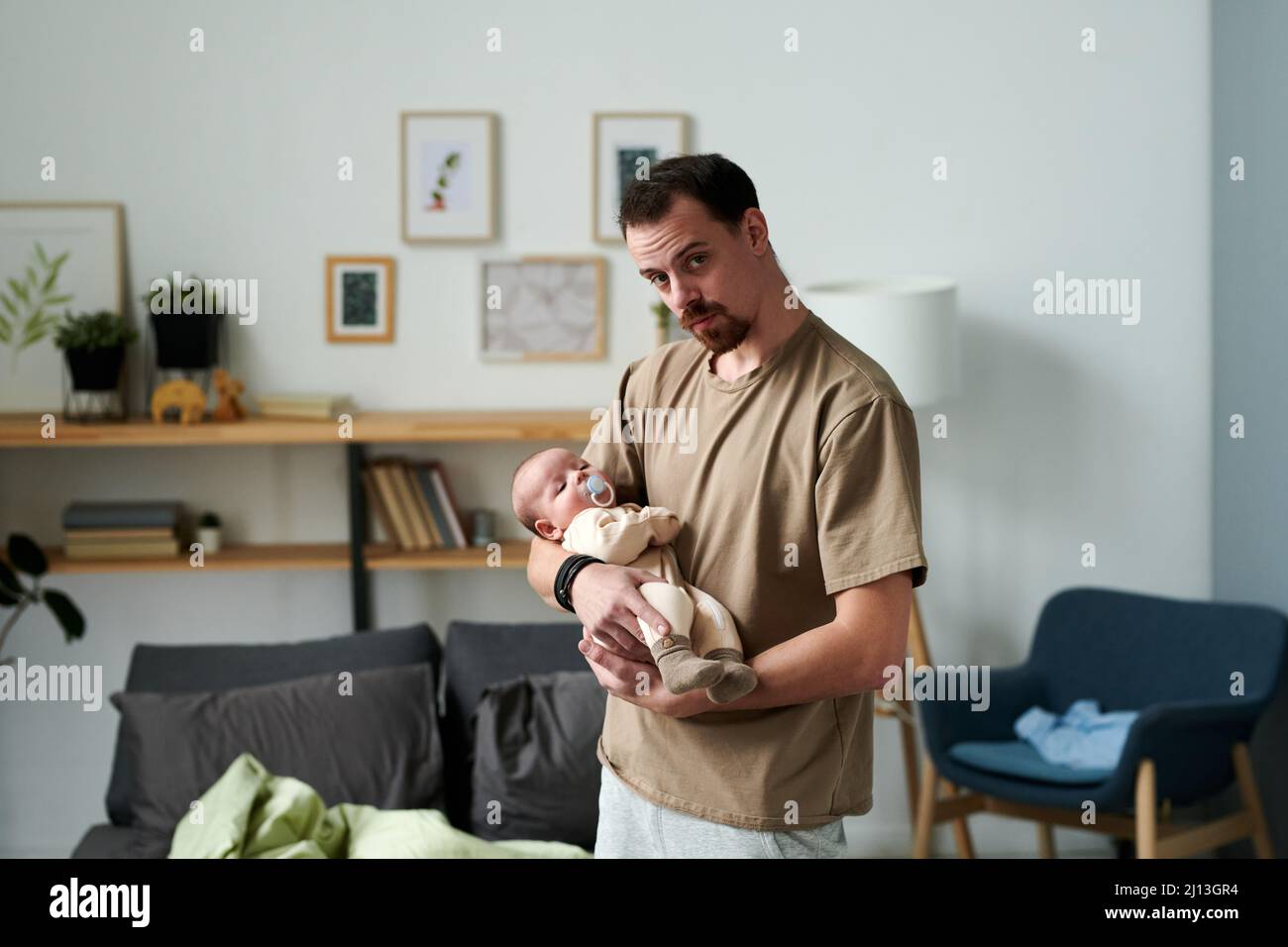 Moderner junger Mann, der seinen kleinen Sohn einlullt, während er vor der Kamera gegen Regale, Bilder in Rahmen, Sessel und Bett steht Stockfoto