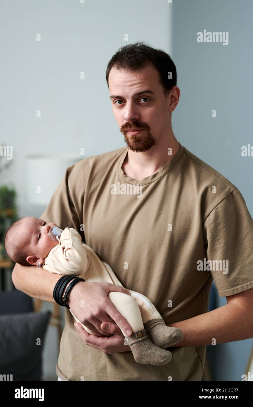 Junger Mann in einem lässigen T-Shirt, der den Jungen in einem Strampelanzug hält, während er vor der Kamera steht und ein Kleinkind einlullt Stockfoto