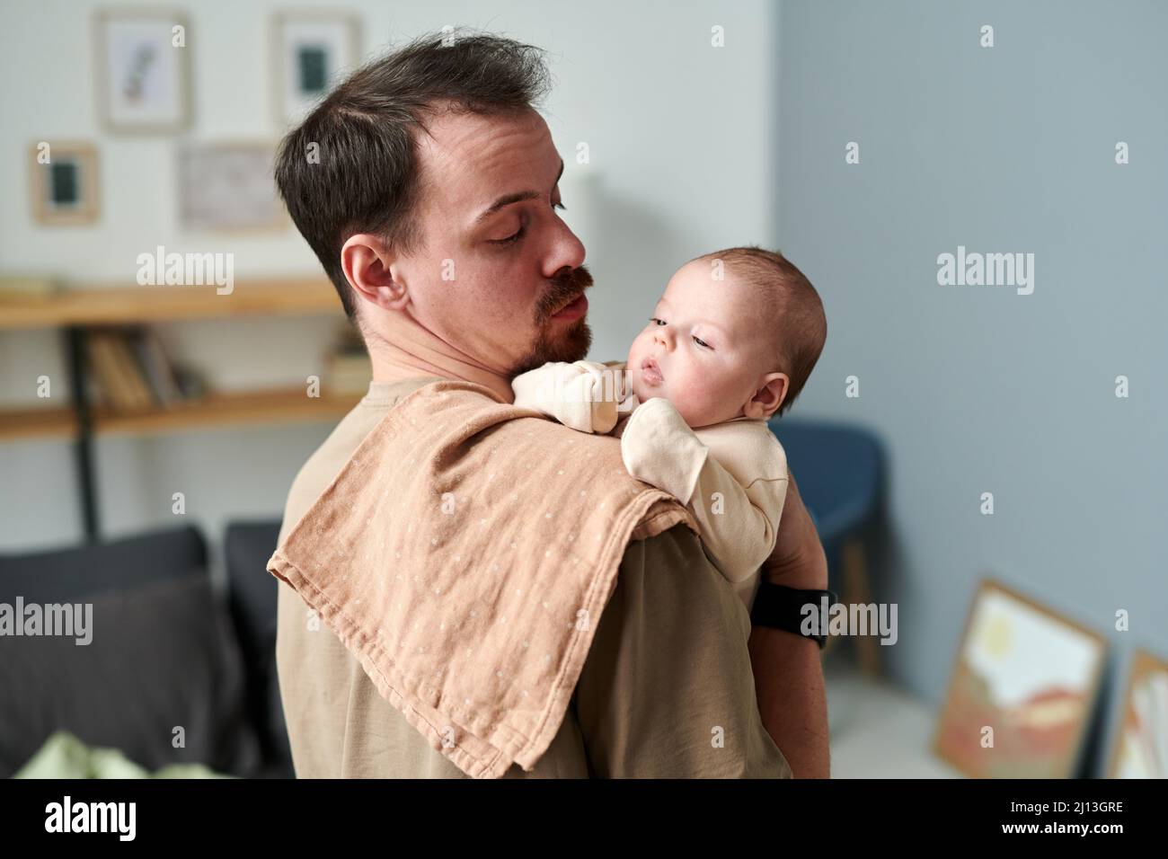 Der junge Vater blickt auf einen entzückenden kleinen Sohn im Strampelanzug auf seiner mit Serviette bedeckten Schulter, während er vor der Kamera steht Stockfoto