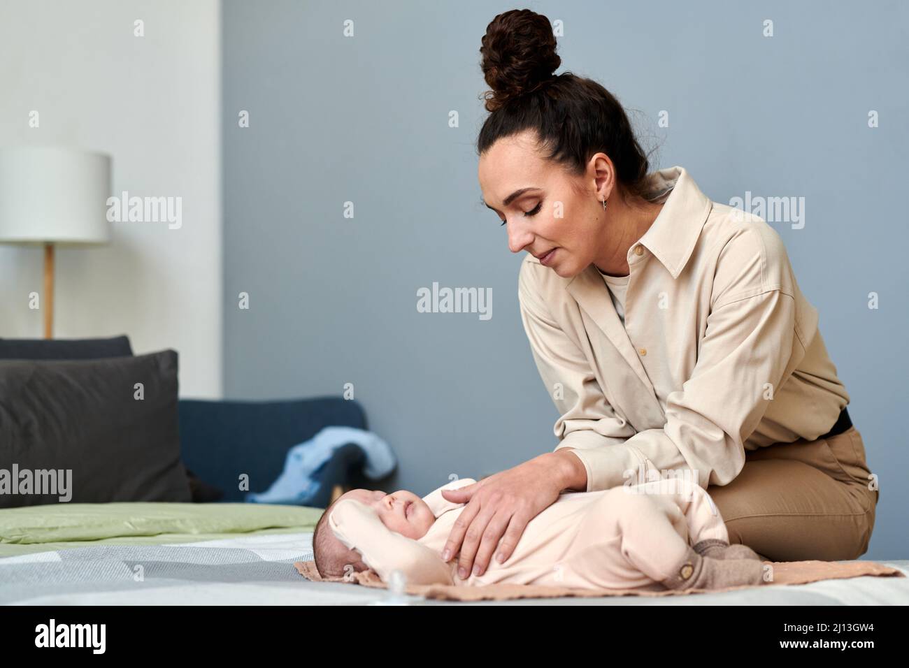 Junge moderne Frau in eleganter Casualwear, die sich über ihren kleinen Sohn beugt, der auf dem Doppelbett liegt, während sie ihn anziehen will Stockfoto