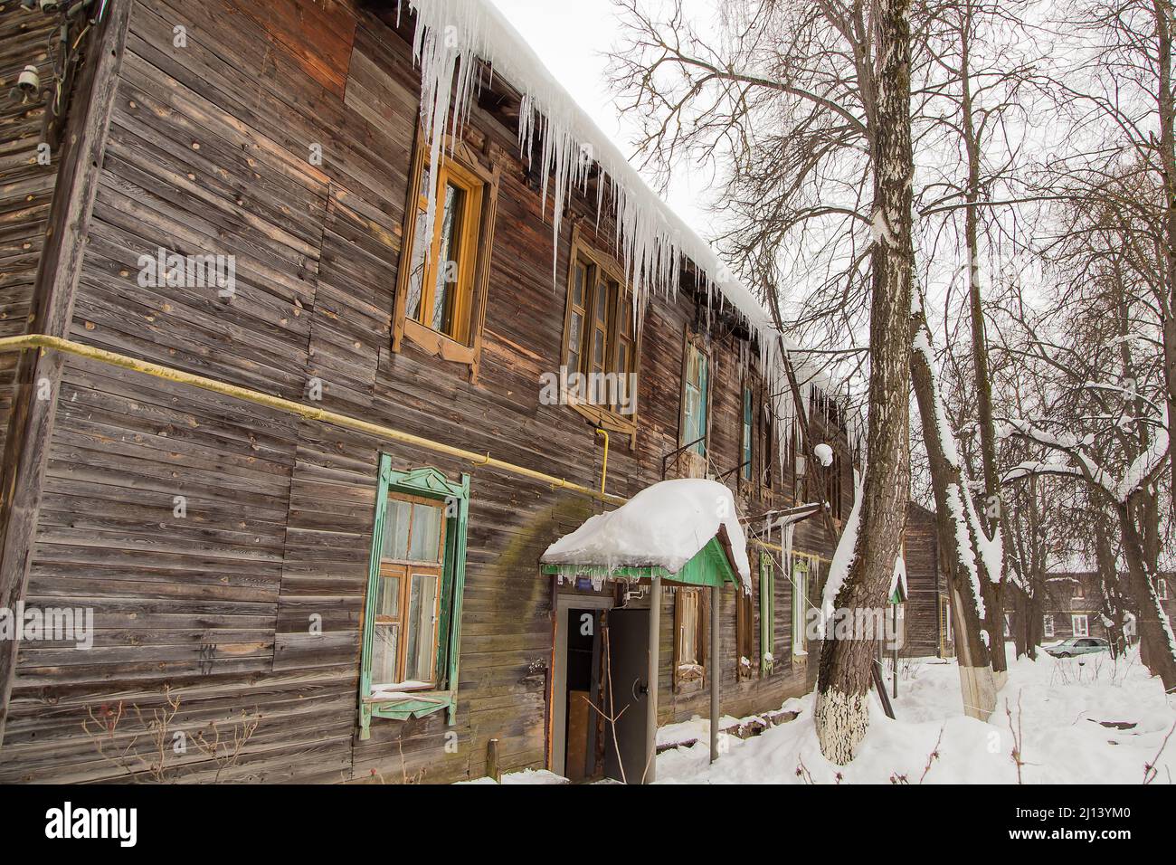 Wachsende transparente Eiszapfen hängen am Rand des Daches. Vor dem Hintergrund der Holzwand des alten Hauses. Große Kaskaden, sogar schöne Reihen. Wolkiger Wintertag, weiches Licht. Stockfoto