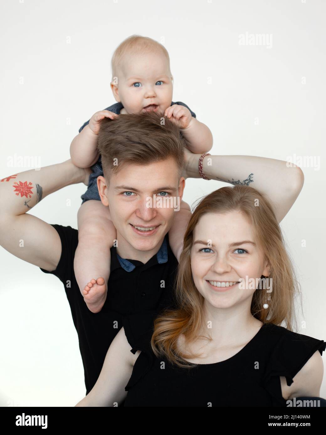 Vertikal lächelnde Familie, die in weißem Studio zusammensteht, Frau, Mann, Baby in schwarzer Kleidung. Kind auf den Schultern halten Stockfoto