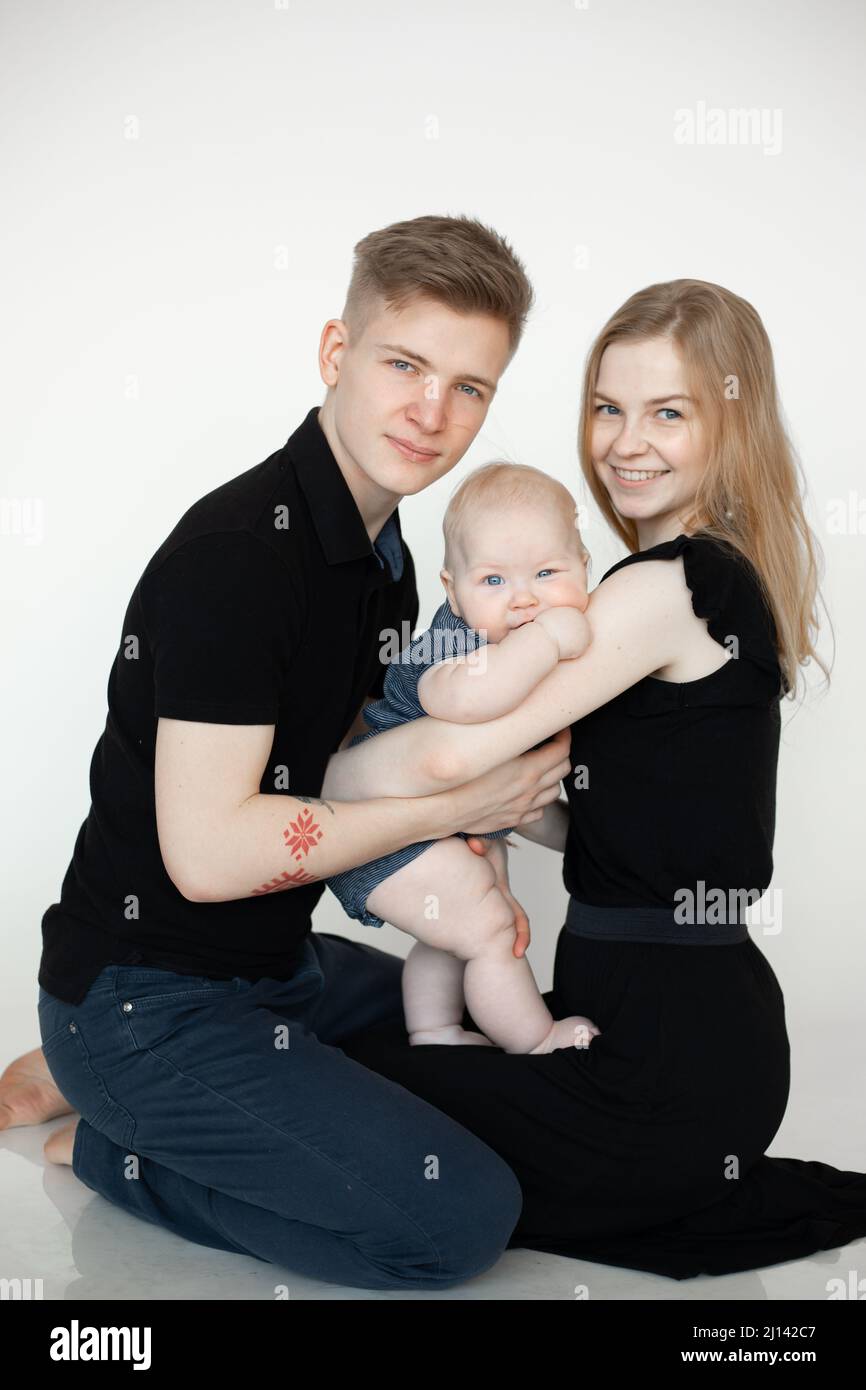 Vertikale glücklich lächelnde blonde Familie sitzt auf dem Knie zusammen, umarmt im Studio. Moderne Familie. Pflege des Neugeborenen Stockfoto