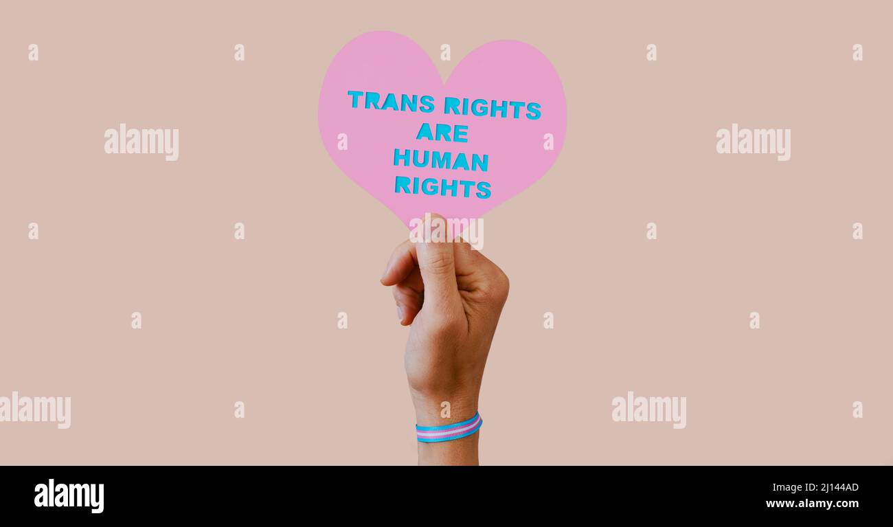 Eine junge kaukasische Person, die ein Armband trägt, das mit der Transgender-Pride-Flagge gemustert ist, hält ein rosa Herz mit dem Text Transgender Rights are human Stockfoto