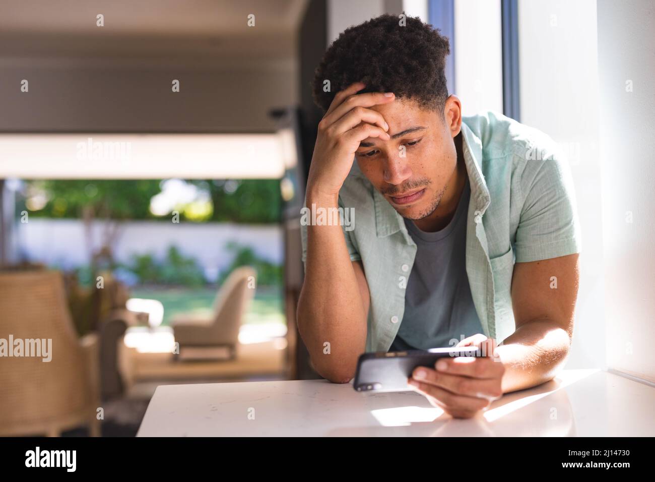Besorgter hispanischer Mann mit Kopf in den Händen starrte auf das Smartphone, das sich zu Hause auf der Küchentheke lehnte Stockfoto