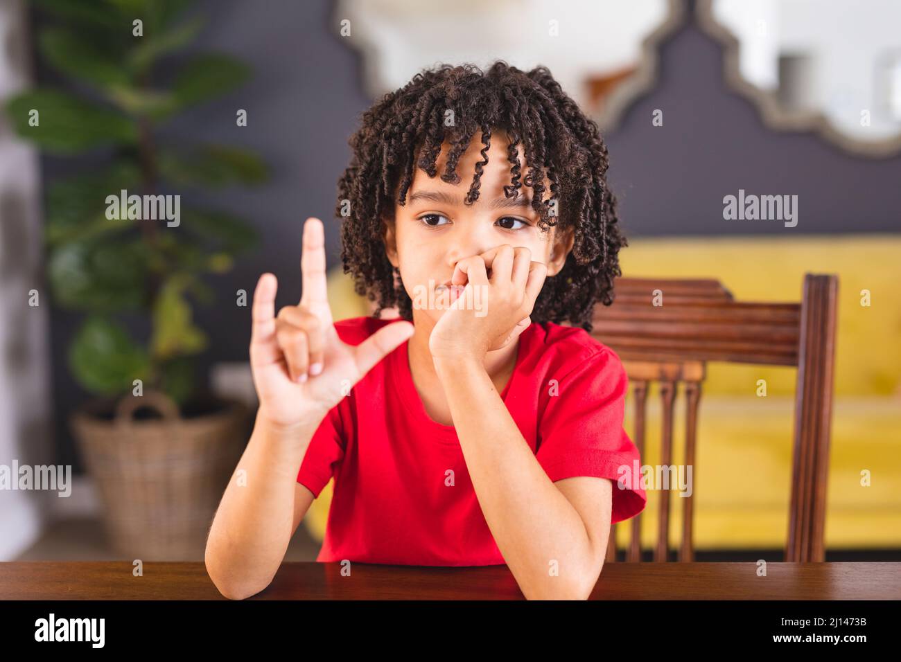 Netter hispanischer stummer Junge, der ein Handzeichen zeigt, während er am Esstisch sitzt Stockfoto