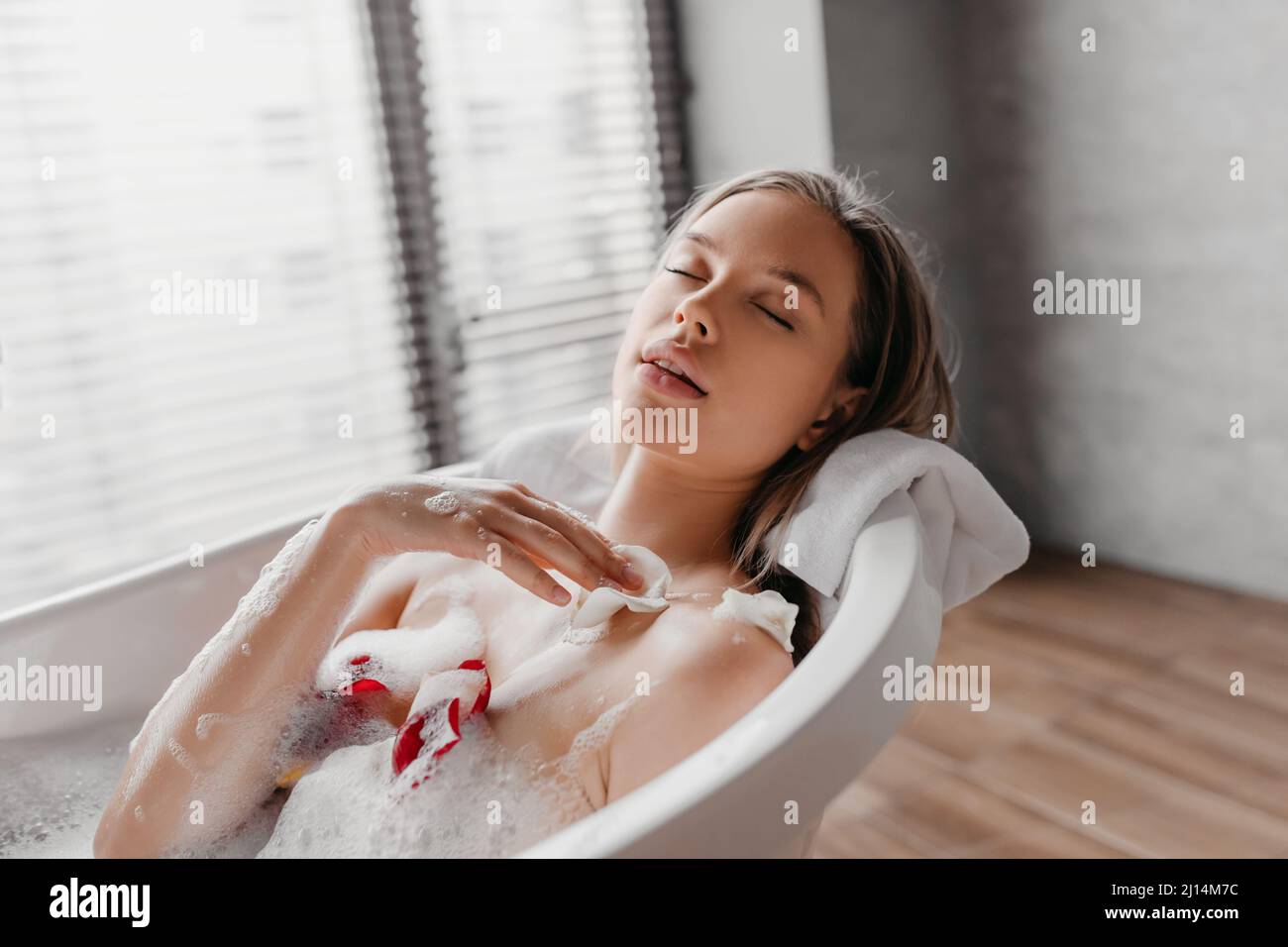 Entspannung und Beauty-Tag zu Hause. Hübsche junge Frau, die in der Badewanne mit Blütenblättern liegt und den heimischen Spa-Tag genießt Stockfoto