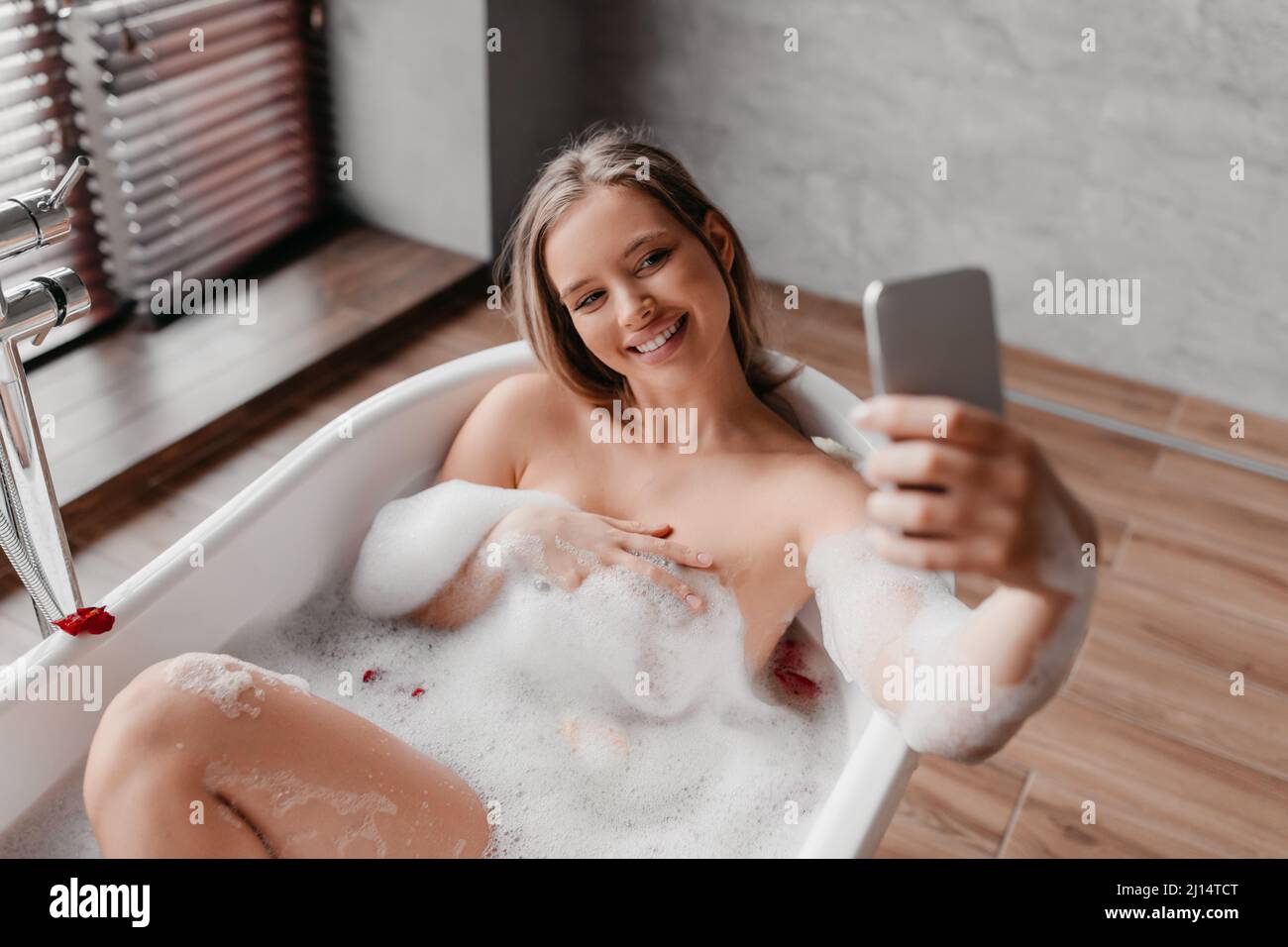 Hübsche Dame, die sich im schäumenden Bad entspannt und Selfie auf dem Smartphone gemacht hat, während sie sich am Morgen an der Schönheitsroutine Vergnügen kann Stockfoto