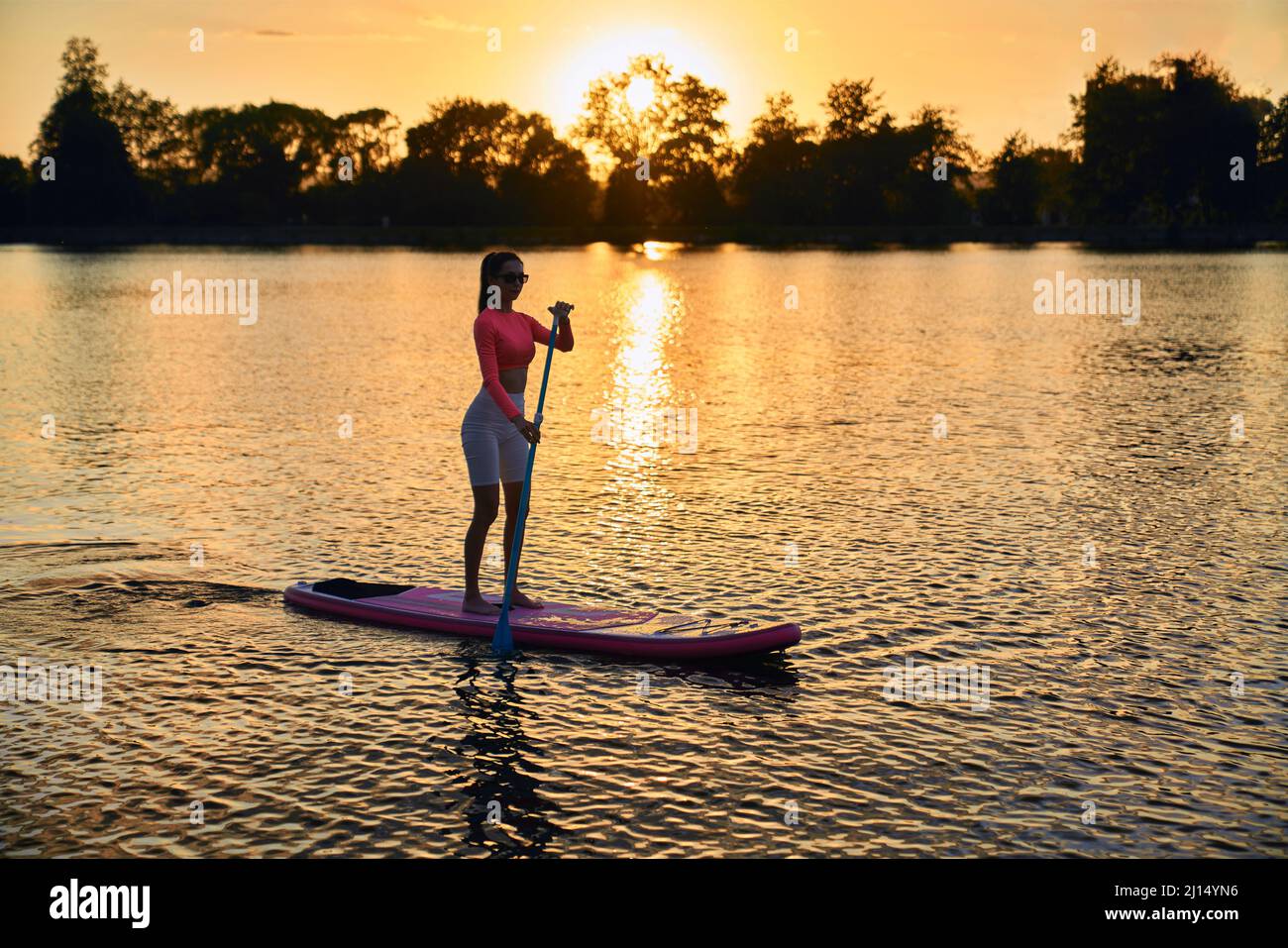 Gesunde starke Frau im Sportoutfit Surfen auf dem Paddle Board während des Sommeruntergangs über dem See. Junge kaukasische Brünette verbringt abends Zeit für Outdoo Stockfoto