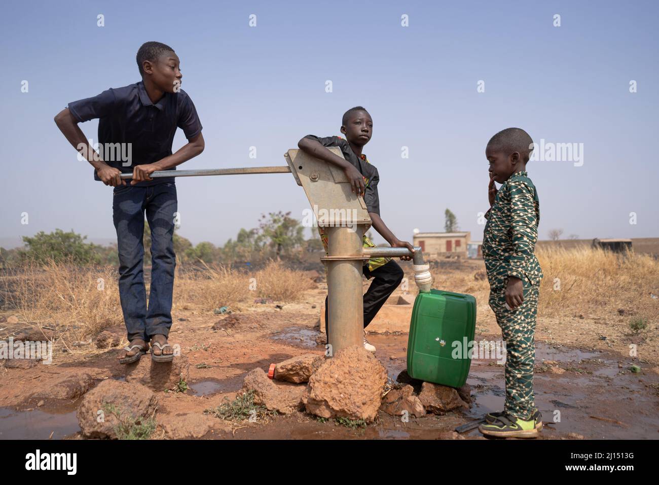 Drei afrikanische Kinder füllen Wasser in einem Kunststofftank an einer Bohrlochpumpe in einer ländlichen Gemeinde in Subsahara-Afrika.Kinderarbeit Konzept. Stockfoto