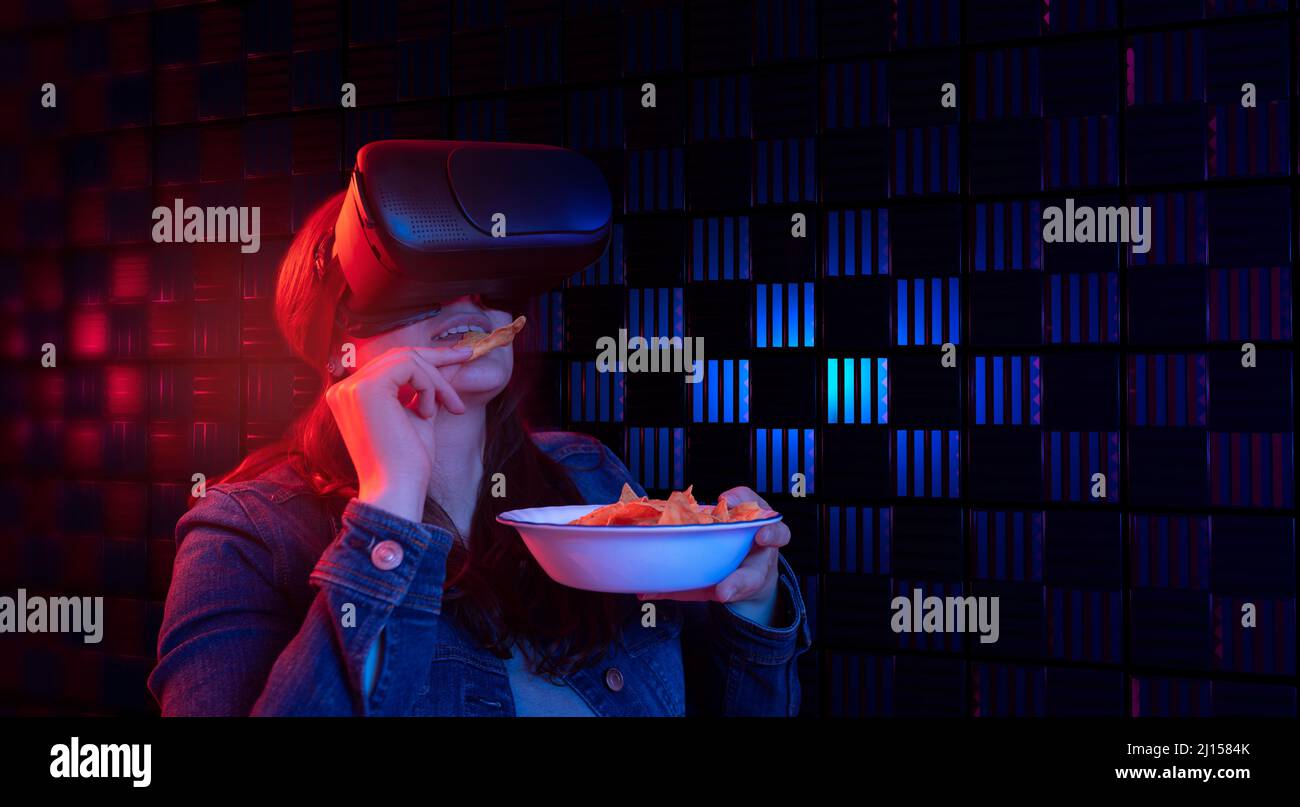 Hispanische Frau mit Virtual-Reality-Brille mit digitaler Technologie, die einen Film aus dem Jahr 3D im Cyberspace mit blauem und rotem Licht beim Essen von Nachos ansieht Stockfoto