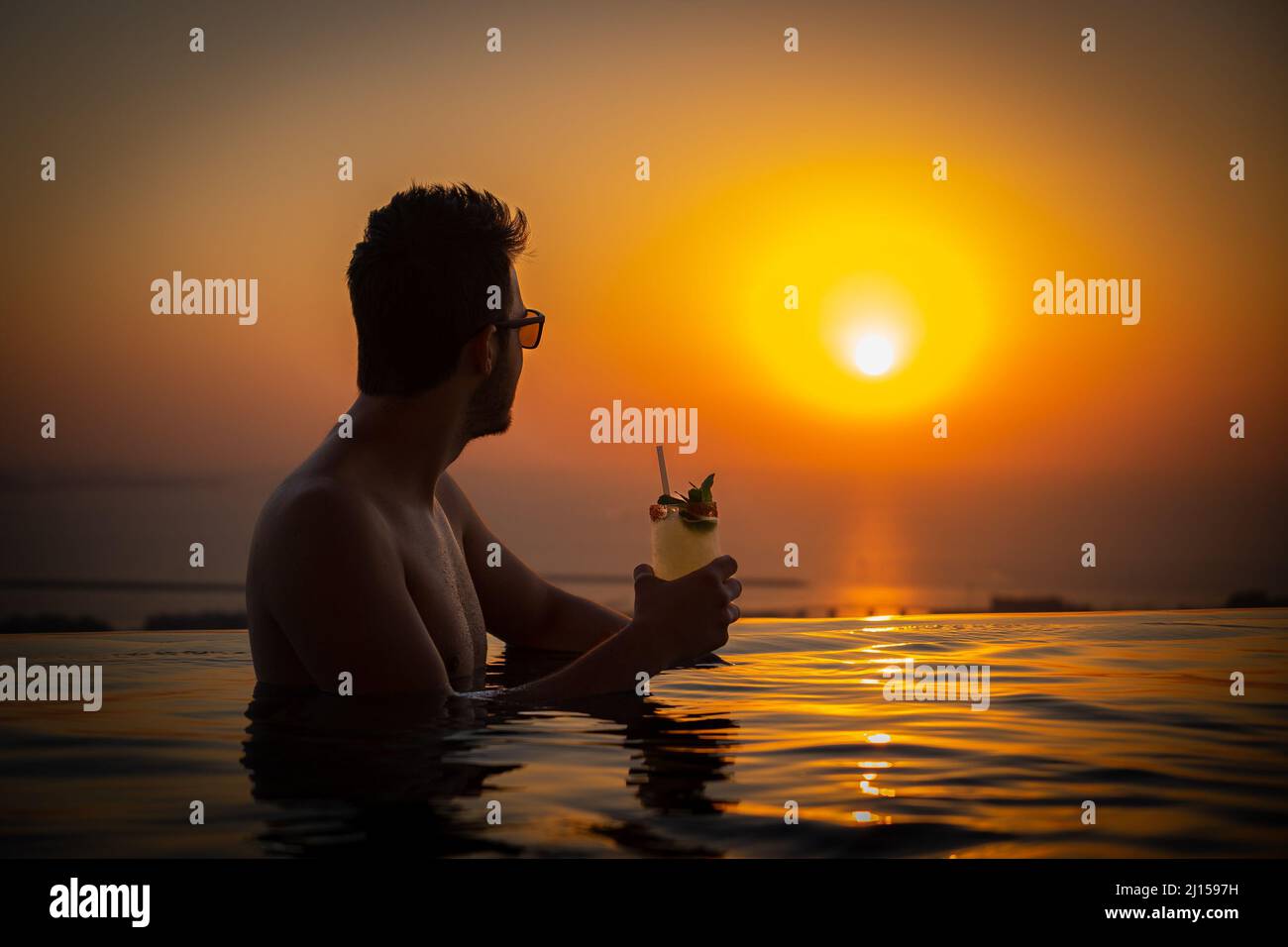 Junger Mann, der in einem unendlichen Pool sitzt und einen Cocktail in der Hand hält, während er das wunderschöne Sonnenuntergangspanorama der Dubai Palm beobachtet Stockfoto
