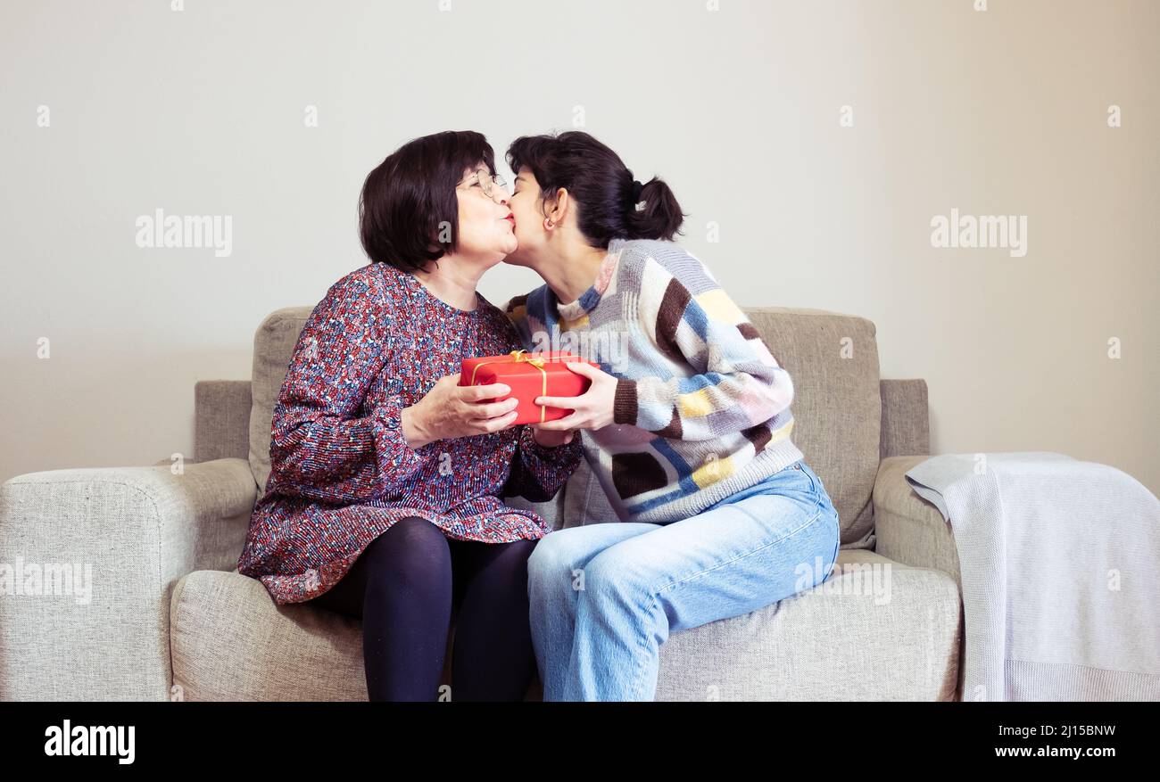 Eine junge Frau umarmt sich und schenkt einer älteren Frau auf ihrer Couch zu Hause ein rotes Geschenk Stockfoto