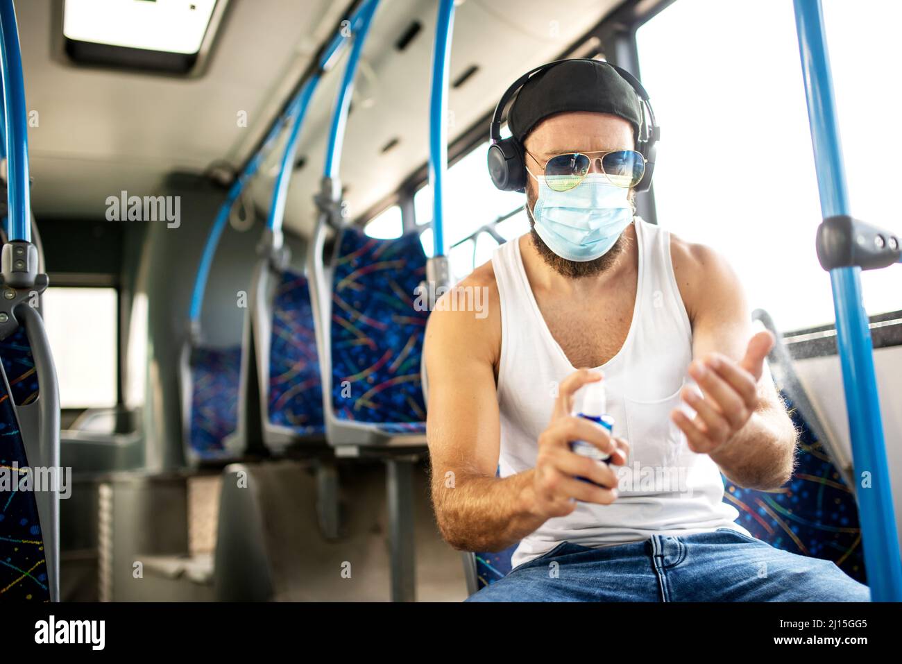 Hübscher blonder Kerl im Unterhemd, der mit öffentlichen Verkehrsmitteln mit Schutzmaske, Sonnenbrille und Kopfhörern reist, respektiert epidemiologische Maßnahmen und Stockfoto