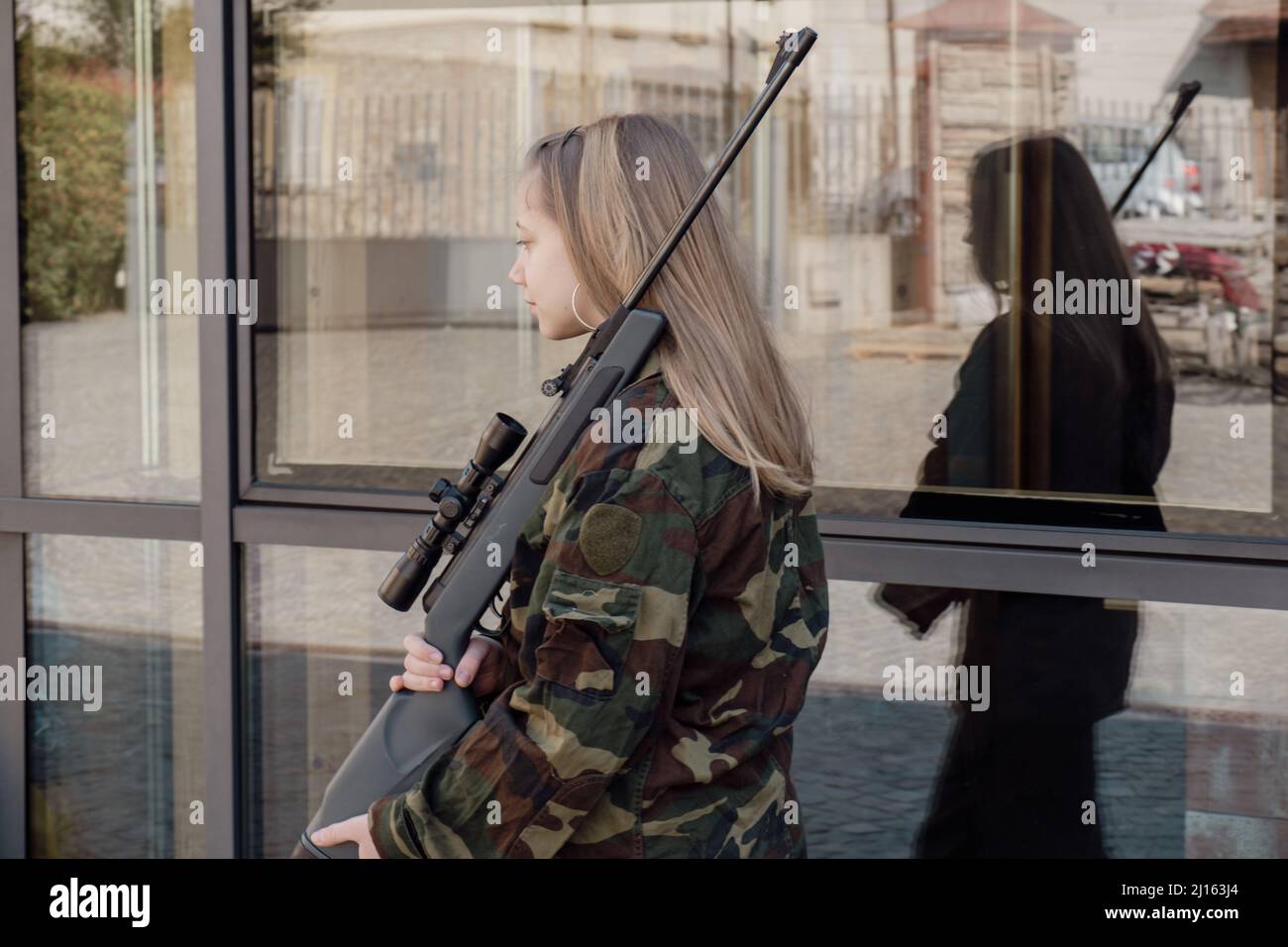 kaukasisch blondes jugendliches Soldatenmädchen an einem Kontrollpunkt mit einem Gewehr und einer camogrünen Militäruniform während eines Konflikts in einem Kriegsgebiet Stockfoto