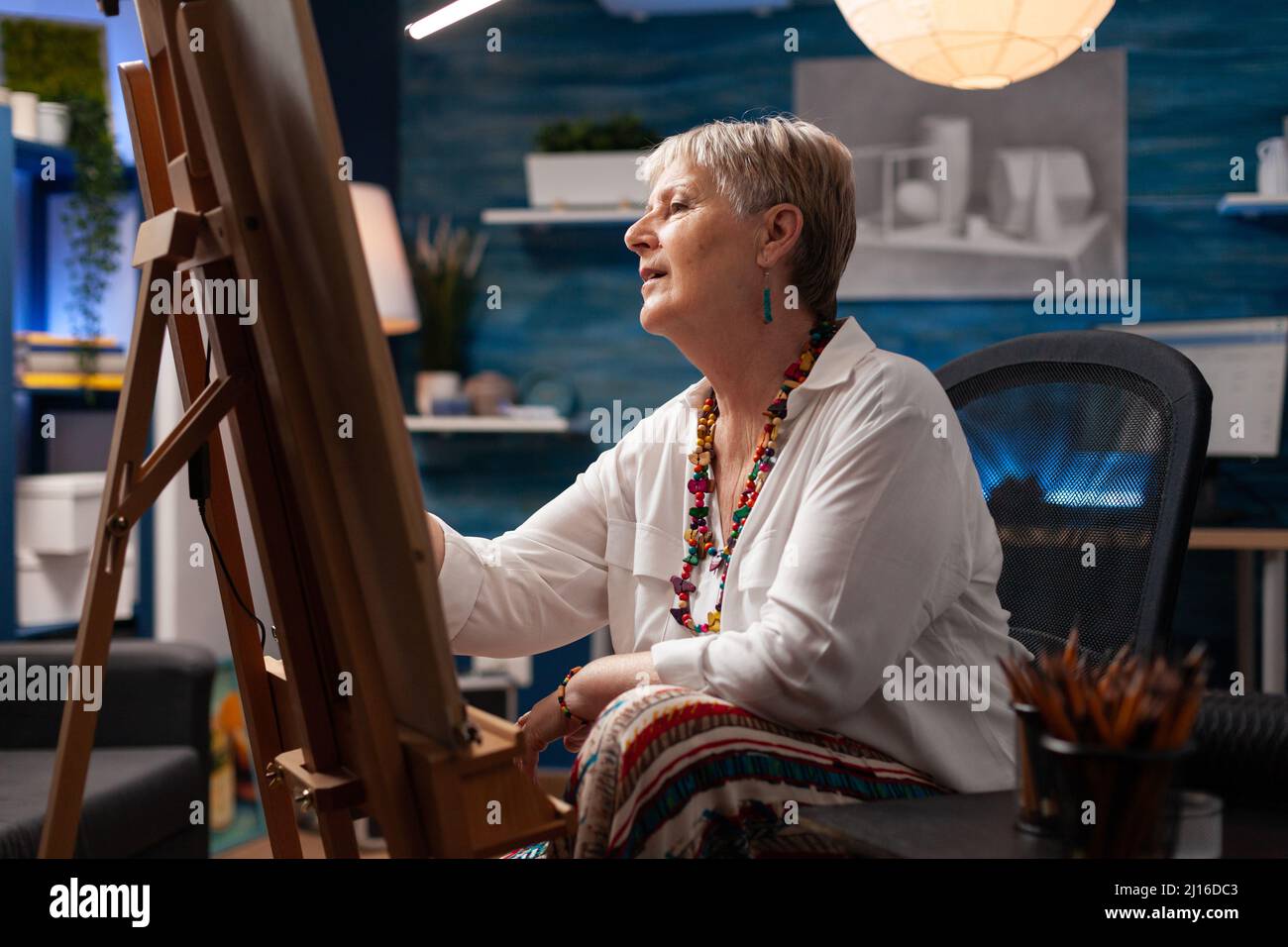 Portrait eines inspirierten Künstlers im Ruhestand vor der Staffelei, der mit Bleistift ein Meisterwerk der kreativen Kunst zeichnet, während er sich hinsetzte. Fokussierte ältere Frau, die im Heimstudio Kohlezeichnungen auf Papier erstellt. Stockfoto