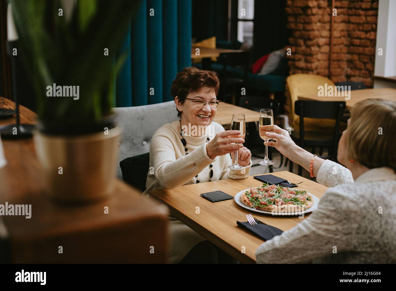 Zwei lächelnde ältere Frauen mit dunklem und hellem Haar, die mit einem Glas Champagner am Tisch sitzen und Pizza feiern. Stockfoto