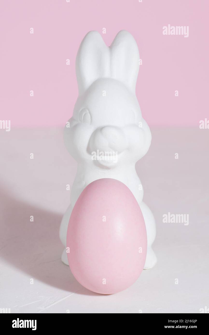 Weißer Osterhase mit einem rosa Osterei auf einem Betontisch auf einem rosa Hintergrund. Harte Schatten. Vertikale Ausrichtung. Speicherplatz kopieren Stockfoto
