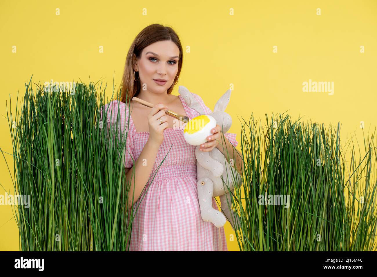 Junge hübsche Schwangerin in rosa, weißem Kleid mit beigefarbenem Spielzeug-Kaninchen und großem künstlichen Ei. Stockfoto