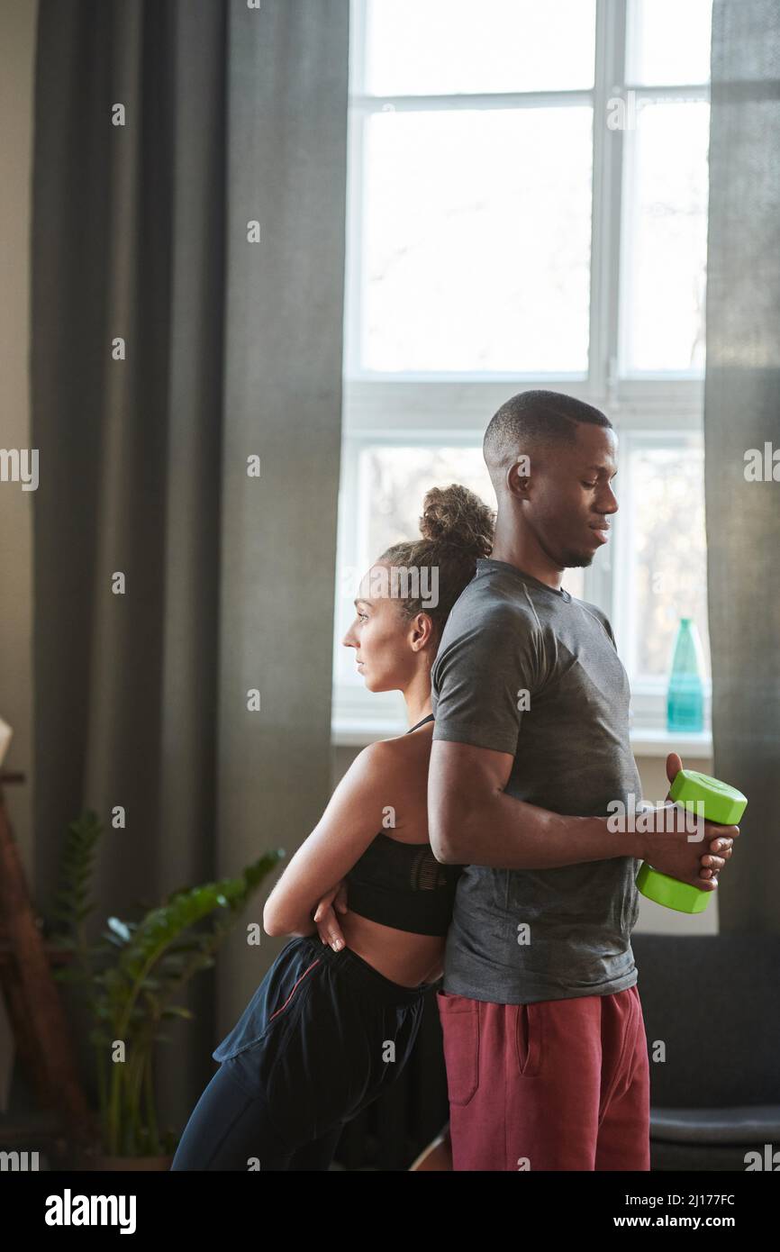 Vertikale Aufnahme des sportlichen jungen schwarzen Mannes und der kaukasischen Frau, die zu Hause im Loft-Wohnzimmer Rücken an Rücken stehen Stockfoto