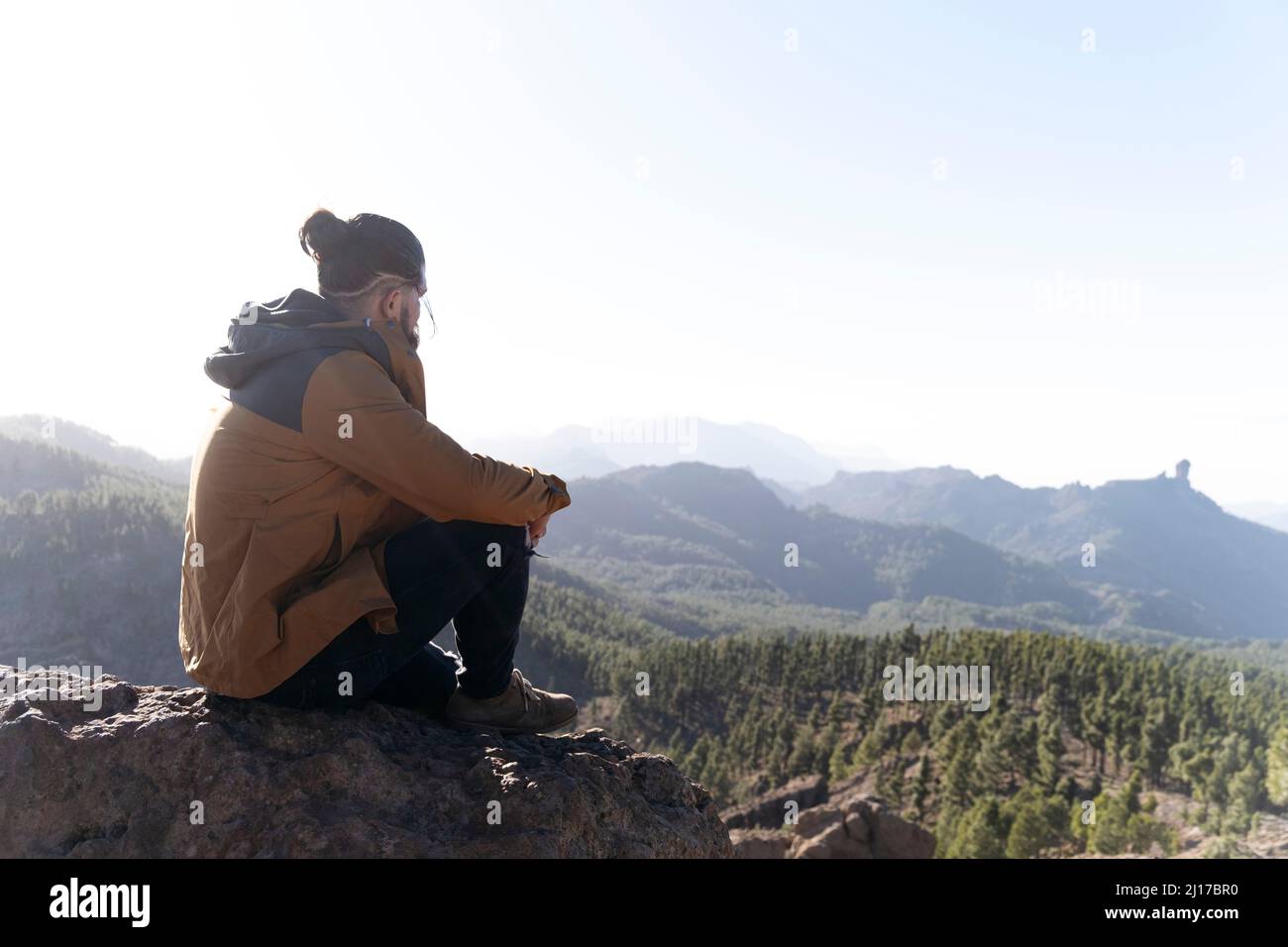 Mann, der auf einem Felsen sitzt und die Aussicht an einem sonnigen Tag betrachtet Stockfoto