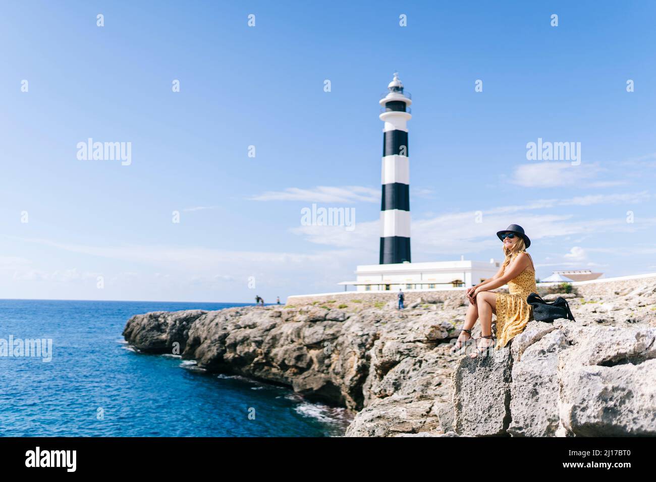 Frau, die am Leuchtturm von Artrutx in Menorca, Spanien, auf Felsen am Meer sitzt Stockfoto