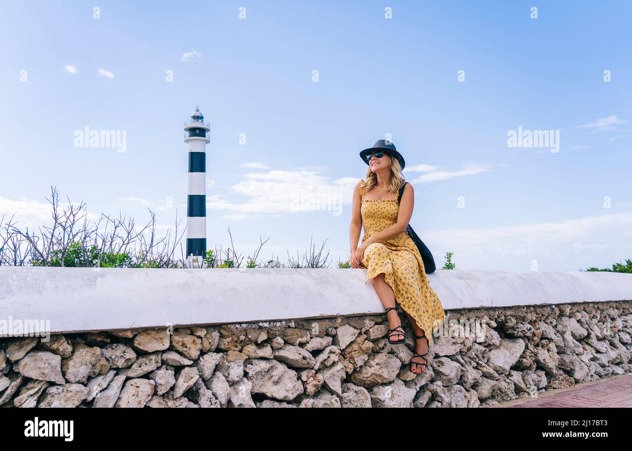 Glückliche Frau, die im Sommer vor dem Leuchtturm von Artrutx auf Menorca, Spanien, sitzt Stockfoto