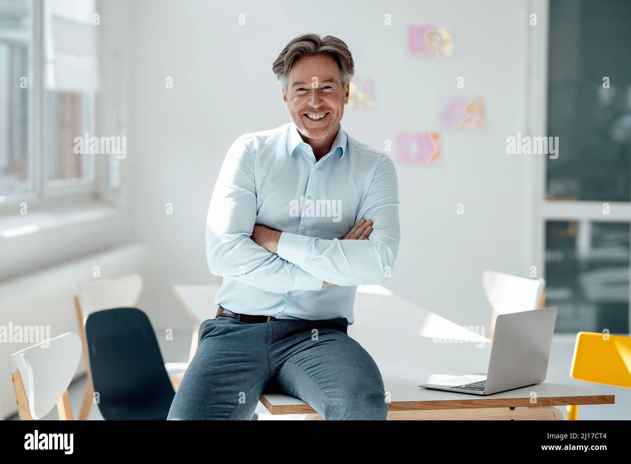 Fröhlicher Geschäftsmann, der mit gekreuzten Armen am Schreibtisch im Büro sitzt Stockfoto