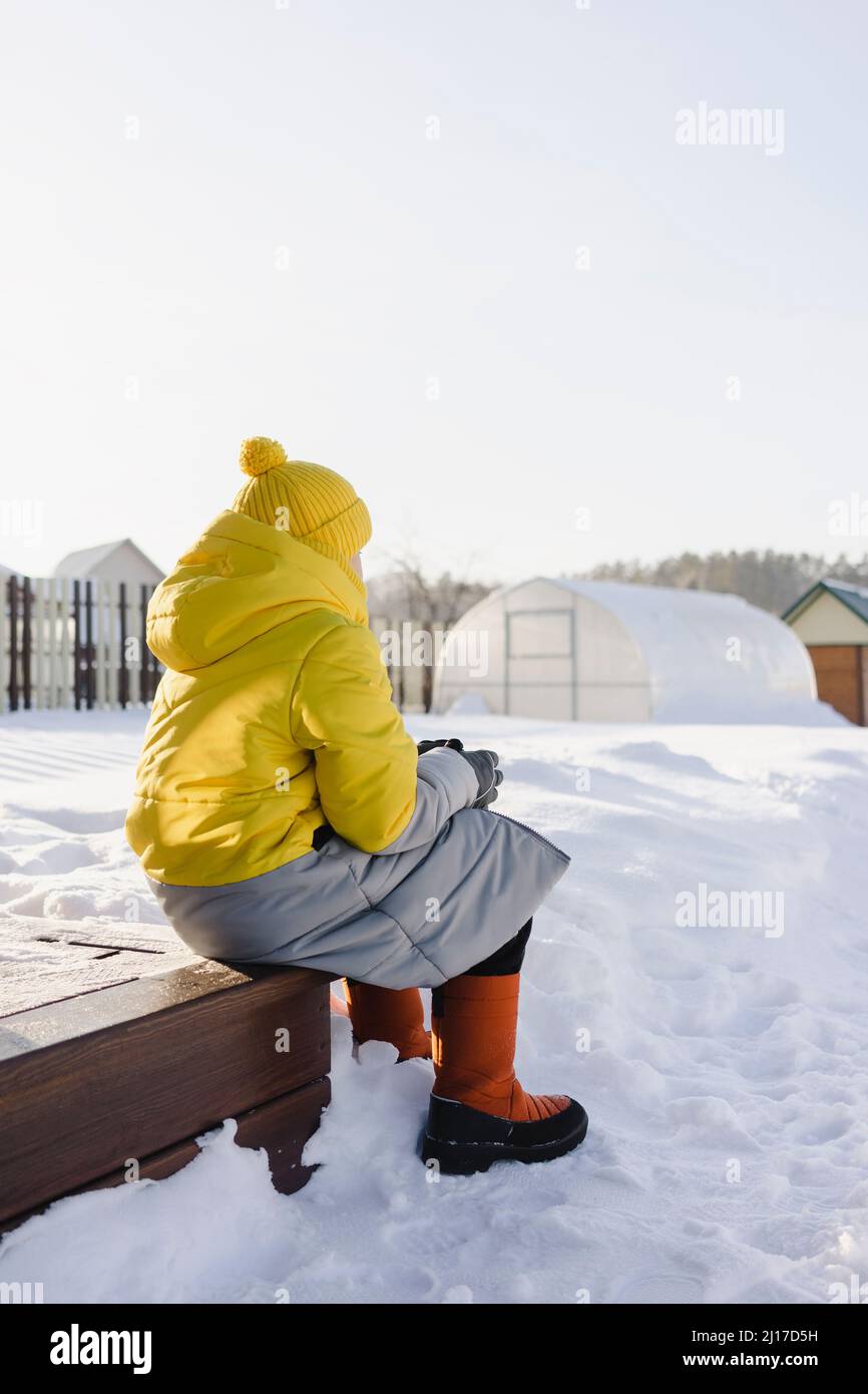 Junge in warmer Kleidung, der im Winter im verschneiten Garten sitzt Stockfoto