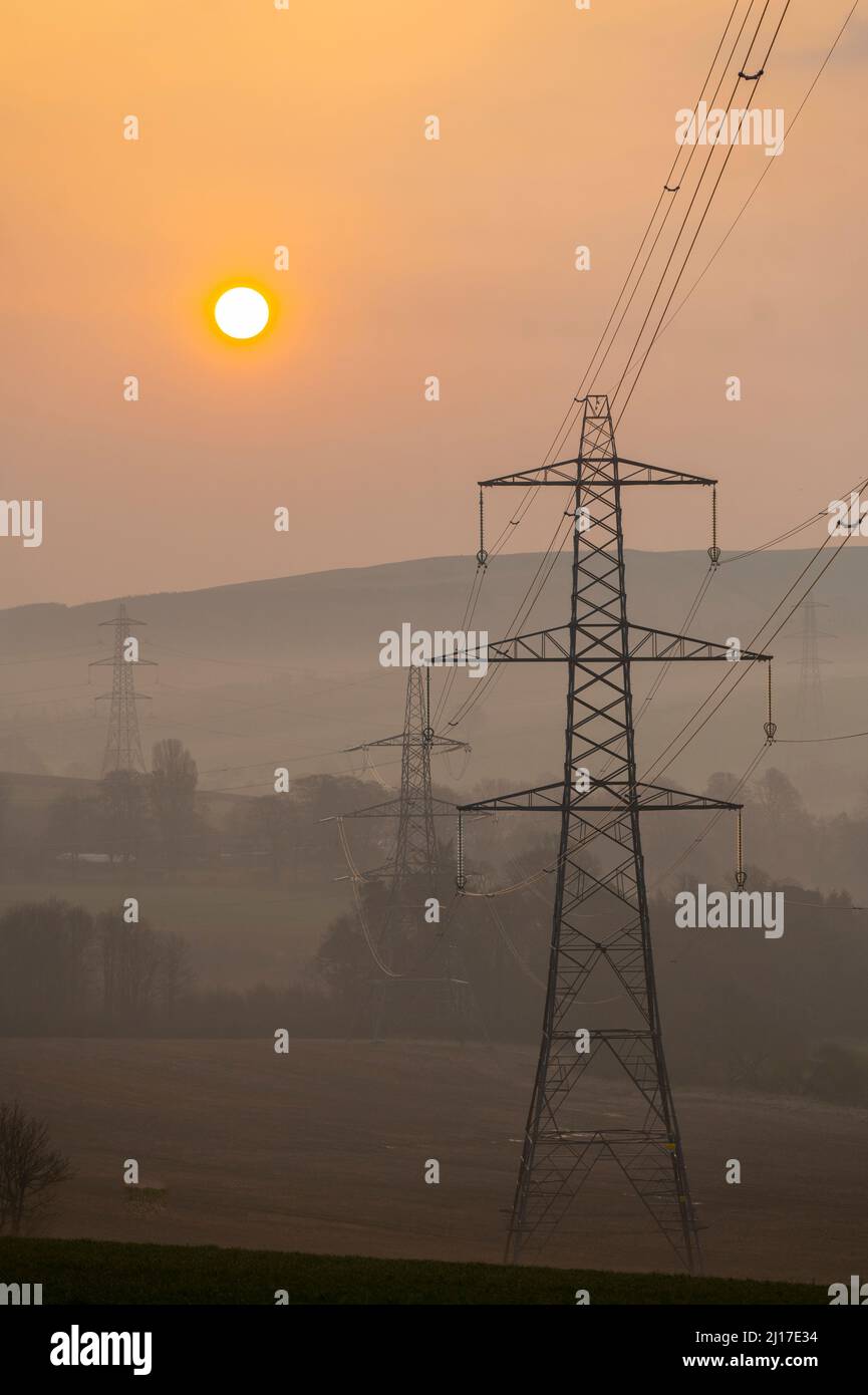 Ledgerwood in Scottish Borders, Großbritannien. 23.. März 2022. Energie, Strom, Strom. Die Sonne geht an einem anderen Tag mit hohen Energiepreisen auf, während die Lebenshaltungskosten weiter steigen. Der Übergang zu grüner Energie steht ganz oben auf der Tagesordnung, aber auch die Bilanz der Senkung der Energiekosten, die derzeit aufgrund der covid Pandemie und des aktuellen Kriegs in der Ukraine und der Sanktionen gegen Russland hoch ist. Der Blick ist auf Strommasten und eine ferne Windturbine, wenn die Sonne in der Nähe von Ledgerwood in den schottischen Grenzen aufgeht. Quelle: phil wilkinson/Alamy Live News Stockfoto