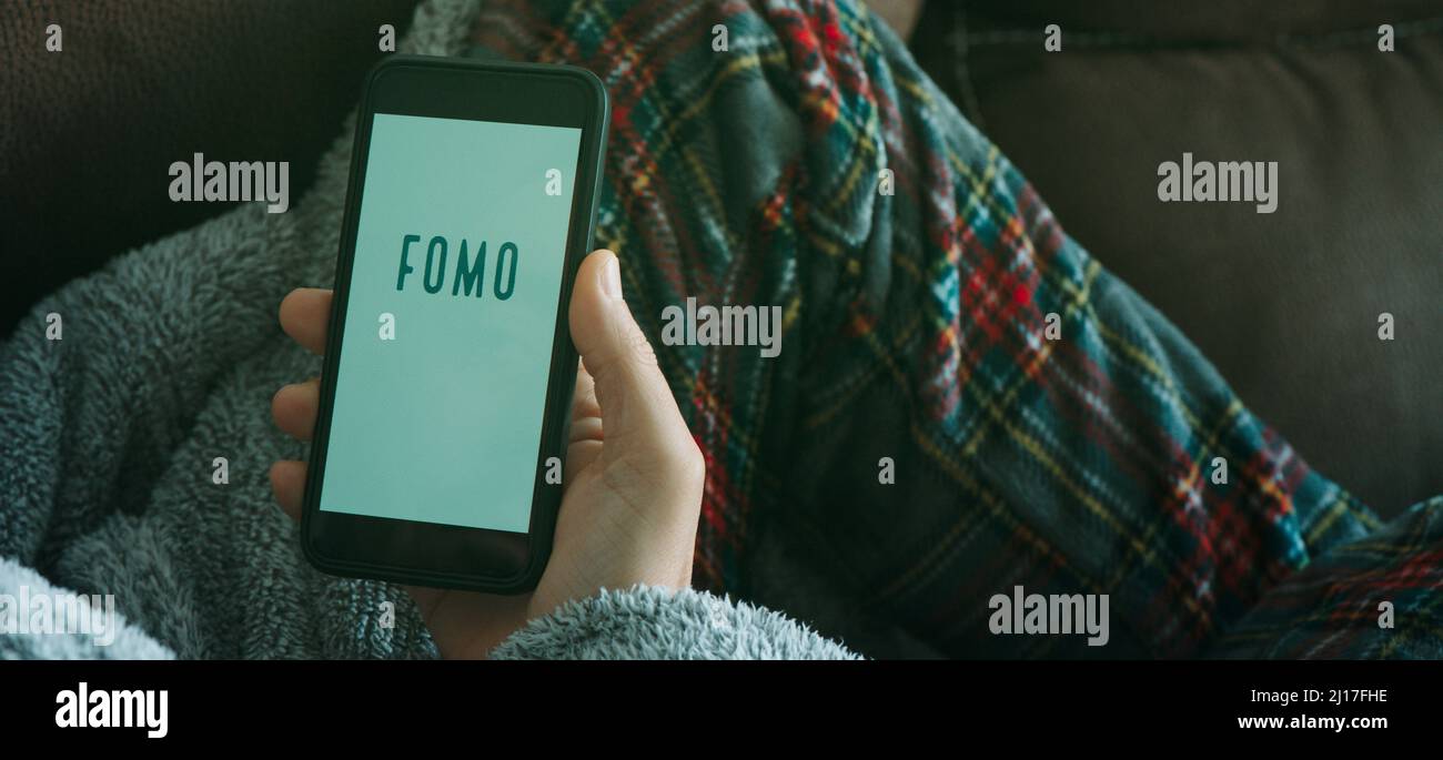 Nahaufnahme eines jungen Mannes auf dem Sofa, der Schlafanzug und Hausmantel trägt und sein Smartphone hält, das den Text fomo aus Angst vor dem Verfehlen in seinem liest Stockfoto