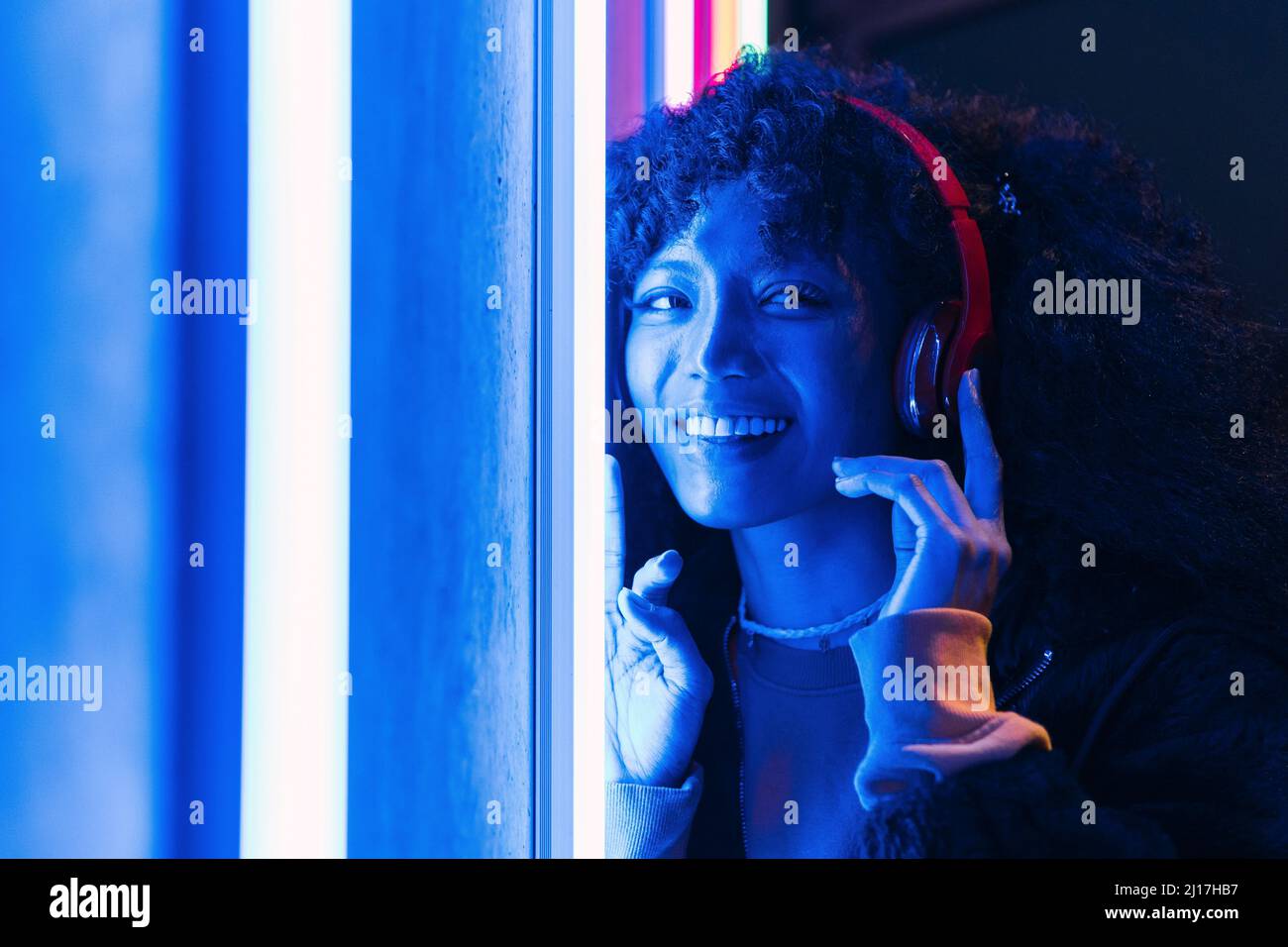 Lächelnde, wunderschöne Frau, die kabellose Kopfhörer an der blau beleuchteten Wand trägt Stockfoto