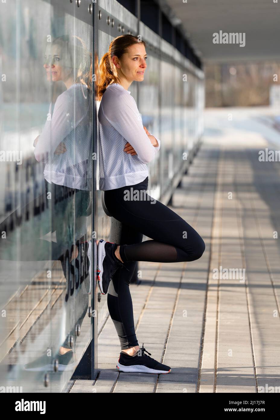 Lächelnde Frau, die mit gekreuzten Armen an einer Glaswand gelehnt steht Stockfoto