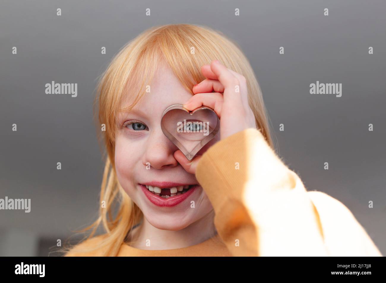 Happy blonde Mädchen hält herzförmige Ausstechform vor dem Auge zu Hause Stockfoto